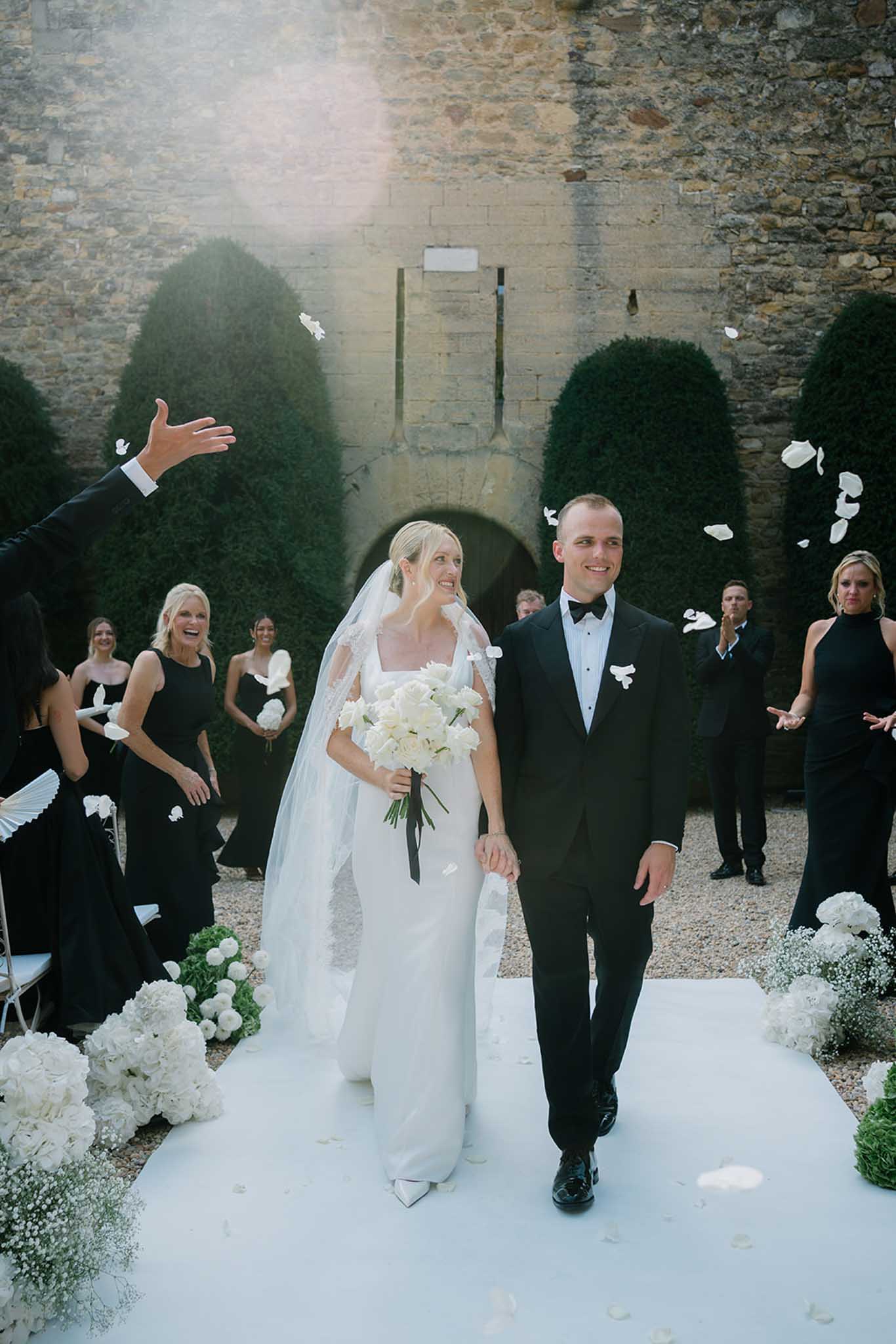 Bride and groom walk white aisle through stone courtyard as guests toss white petals, hydrangeas lining path