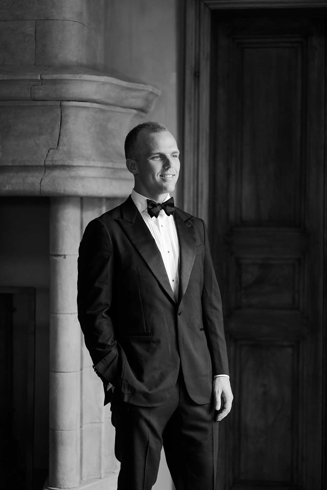 Black-and-white groom portrait in black tuxedo standing beside stone column and paneled door in elegant interior