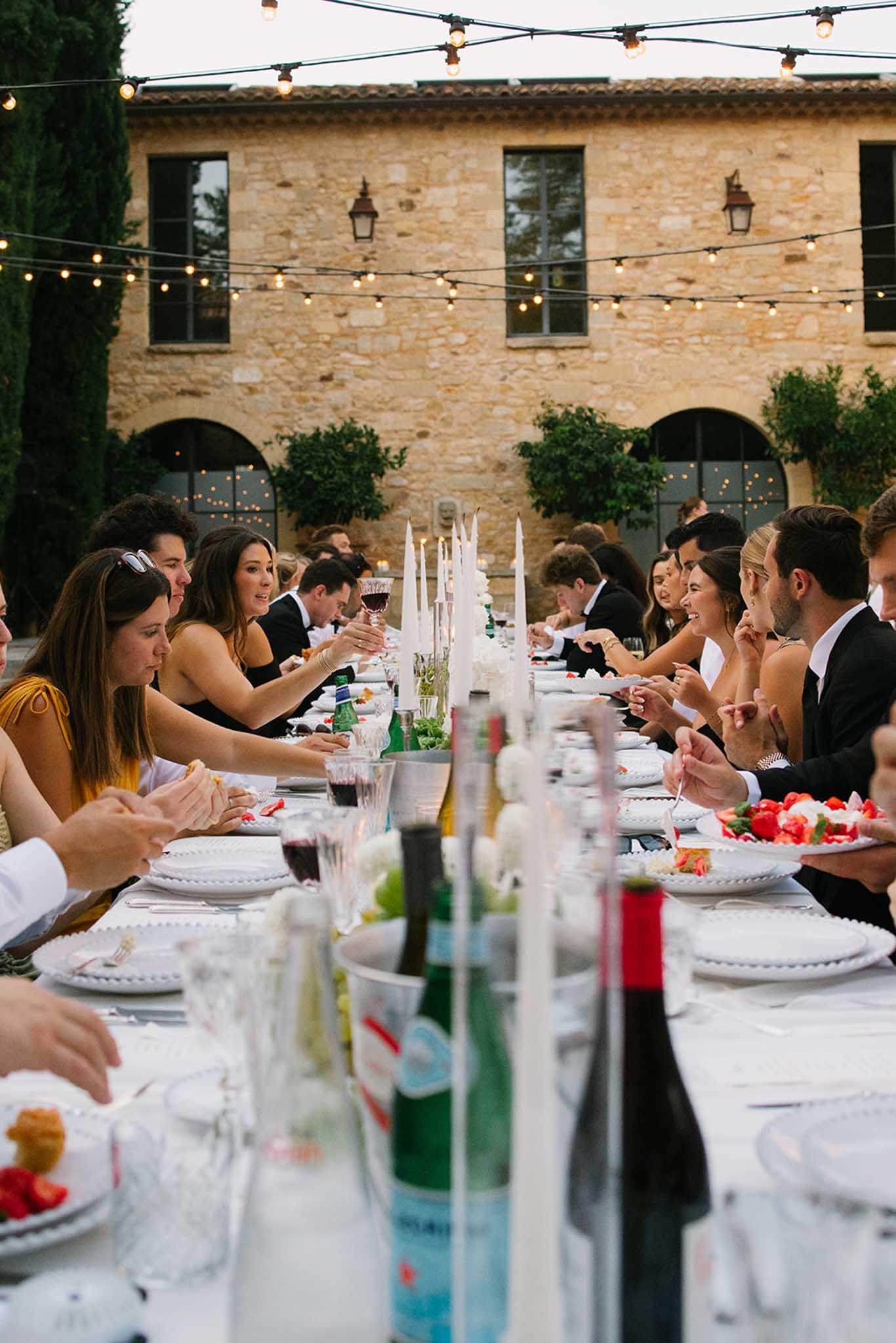 Guests seated at a long reception table in a stone courtyard with string lights, taper candles, and floral arrangements