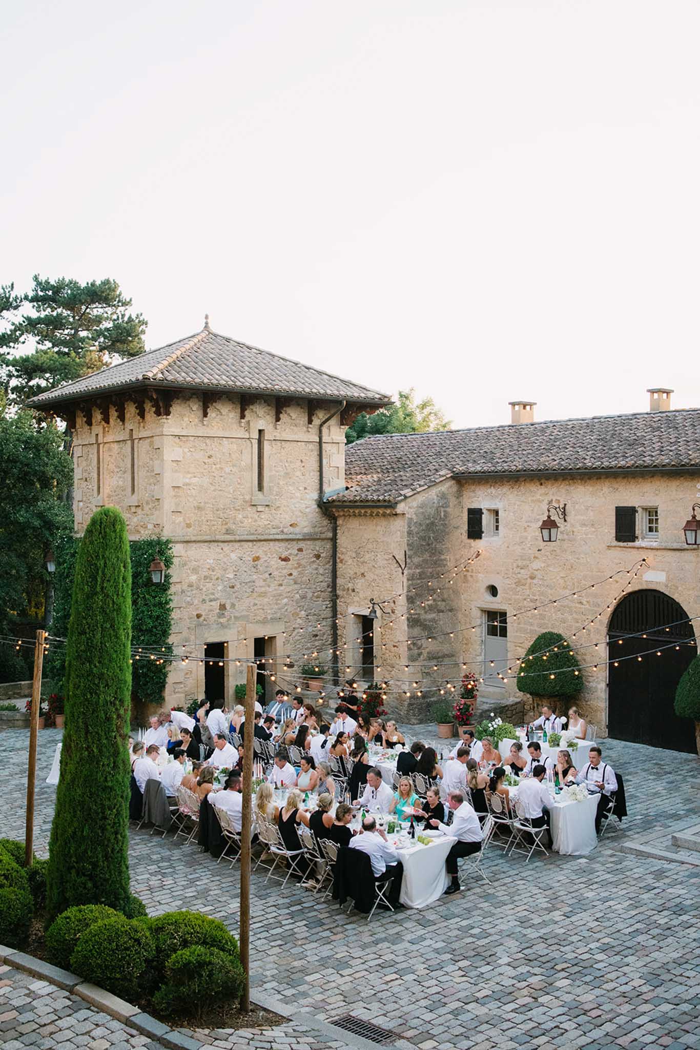 Wedding reception in a historic stone chÃ¢teau courtyard with round tables, string lights and cylindrical cypress trees; circular tower on left.