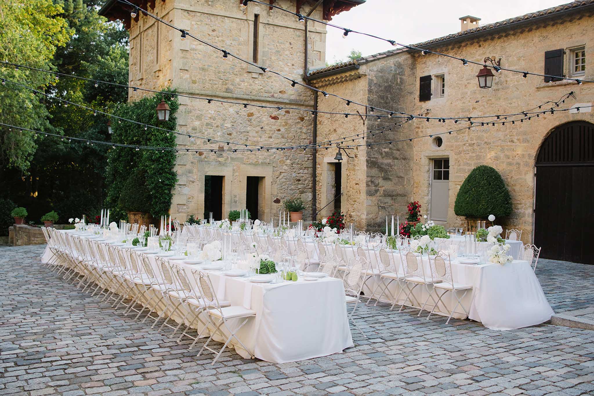 Wedding reception tables with white linens and green floral centerpieces in Tuscan stone courtyard with string lights overhead
