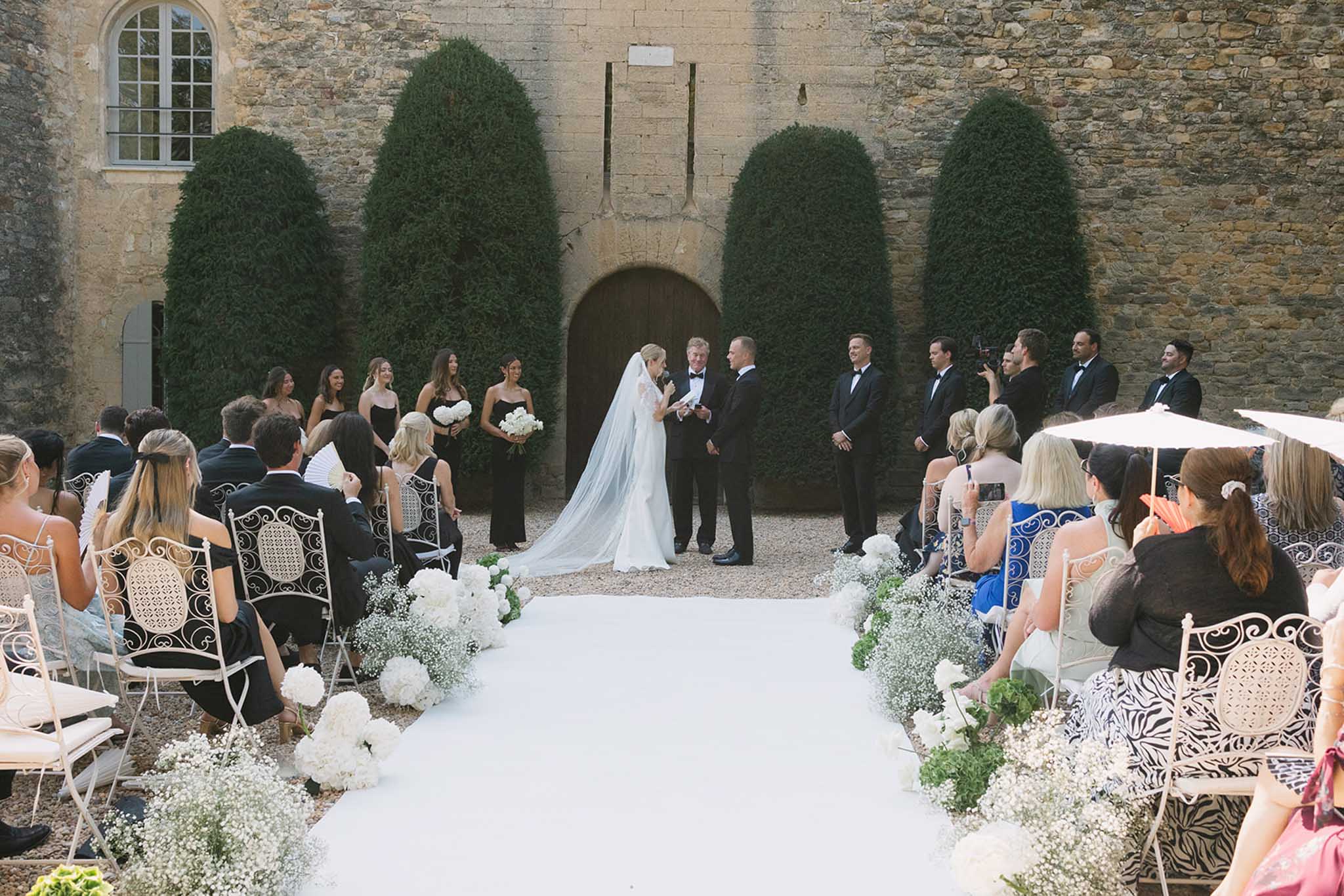 Outdoor ceremony in stone courtyard with white hydrangea aisle markers and manicured topiaries framing altar