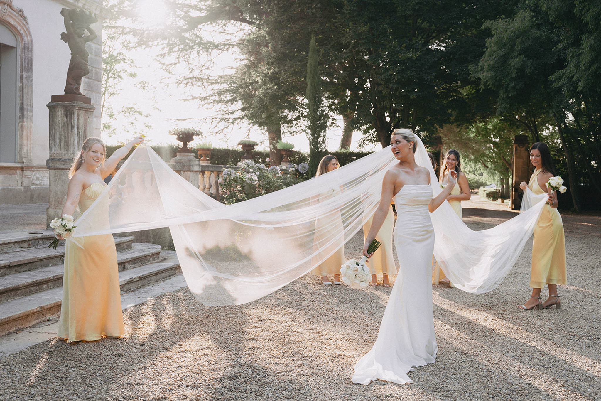 Bride with billowing veil and bridesmaids in pastel dresses in garden courtyard