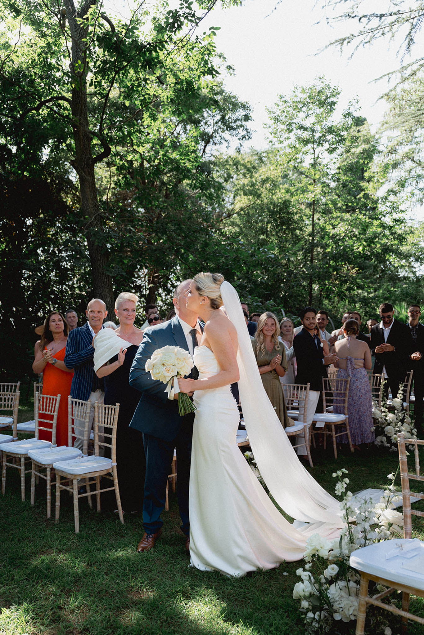 Outdoor wedding ceremony under mature trees with guests seated in white chiavari chairs and white hydrangea aisle flowers