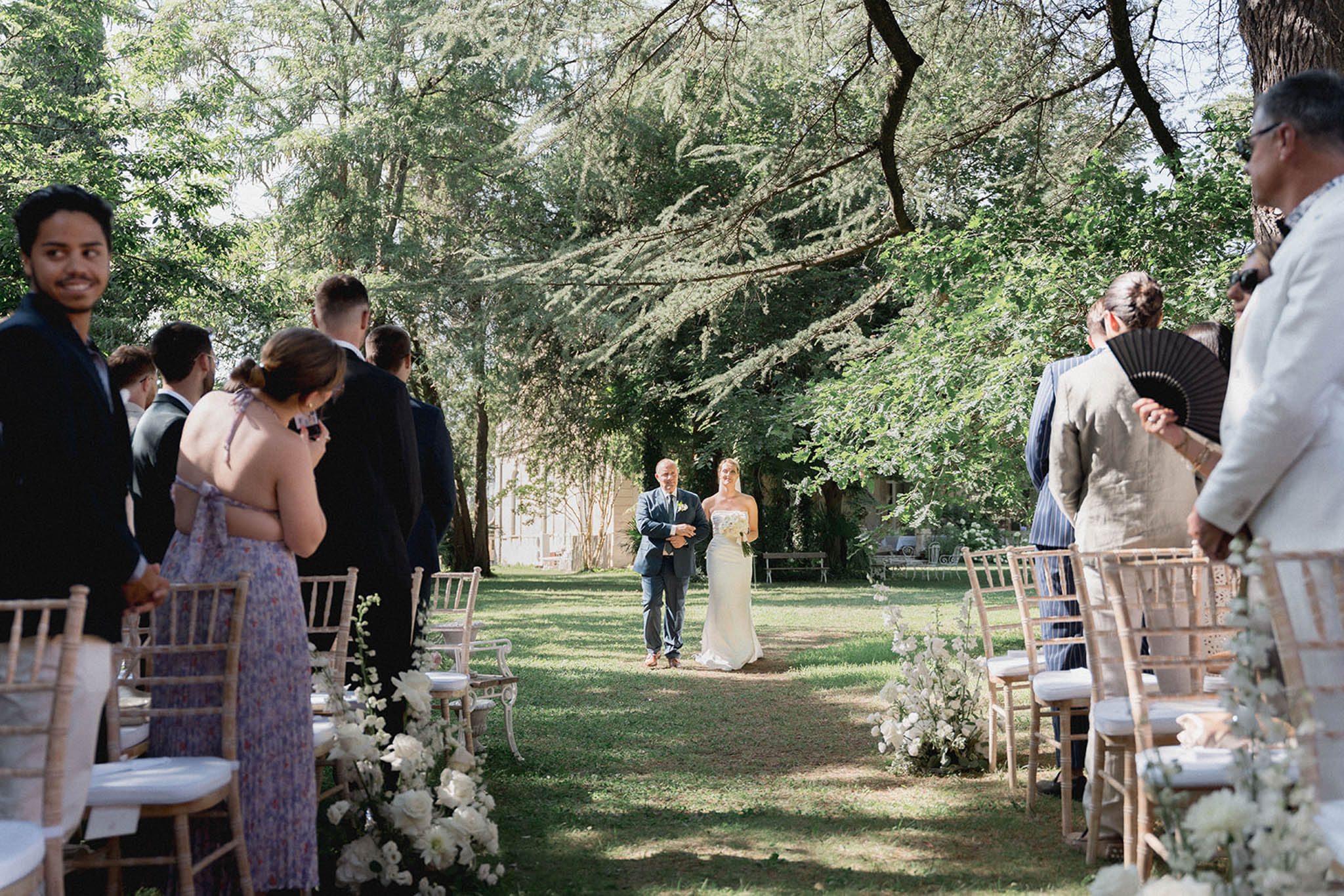 Father of the bride walking daughter down garden ceremony aisle lined with white roses under mature trees