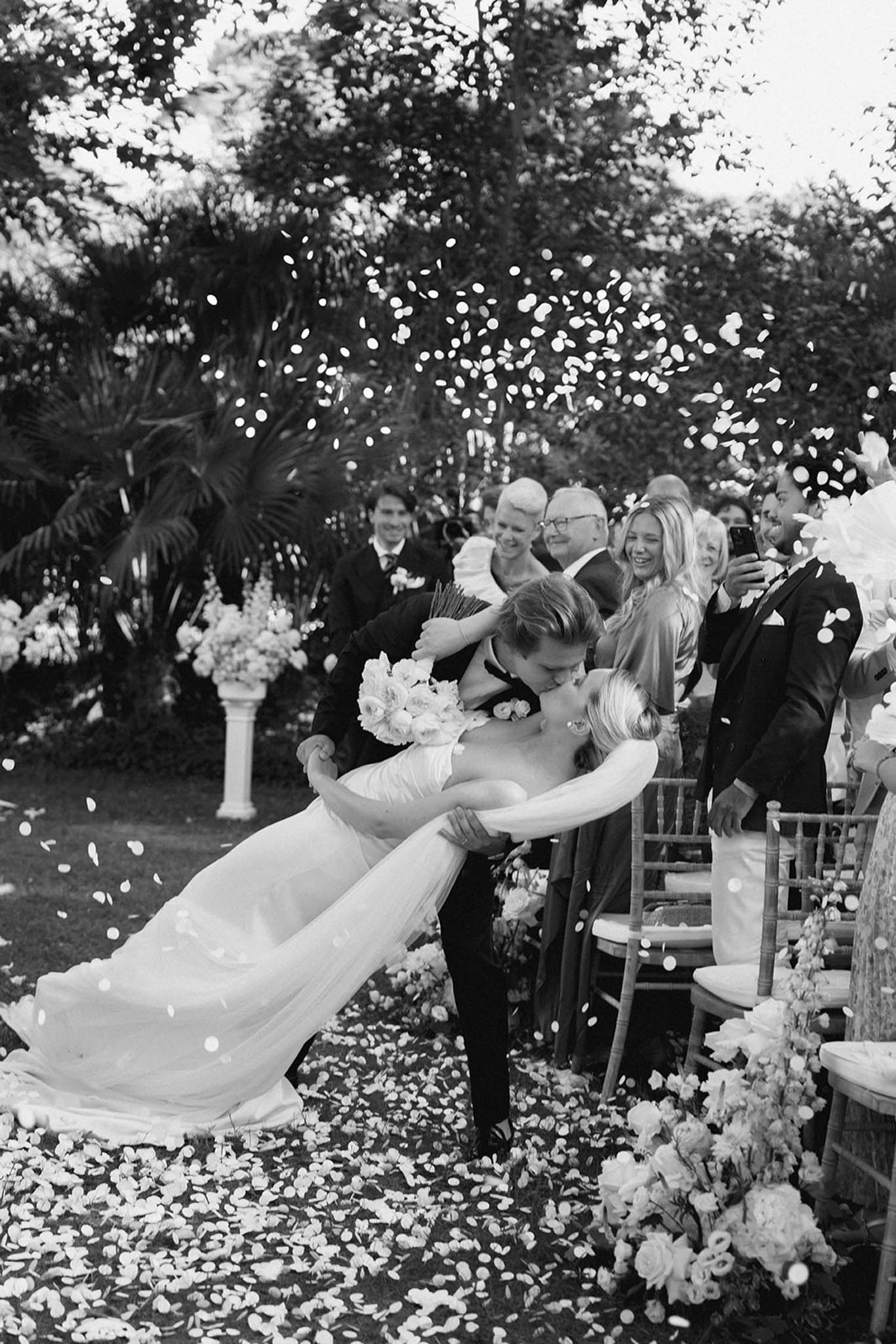 Black-and-white groom dipping bride on petal-strewn aisle during recessional