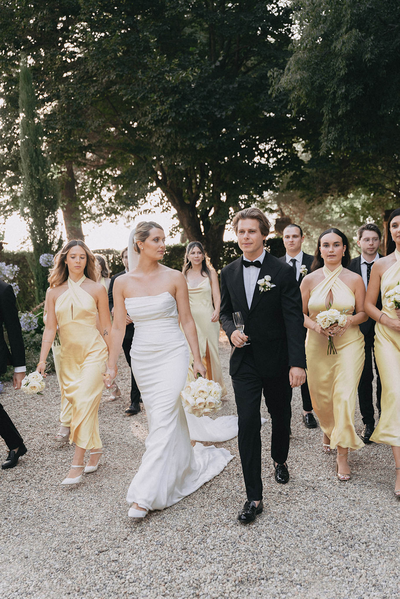 Bride and groom in recessional on tree-lined path, bridesmaids in champagne dresses, groomsmen in black tuxedos
