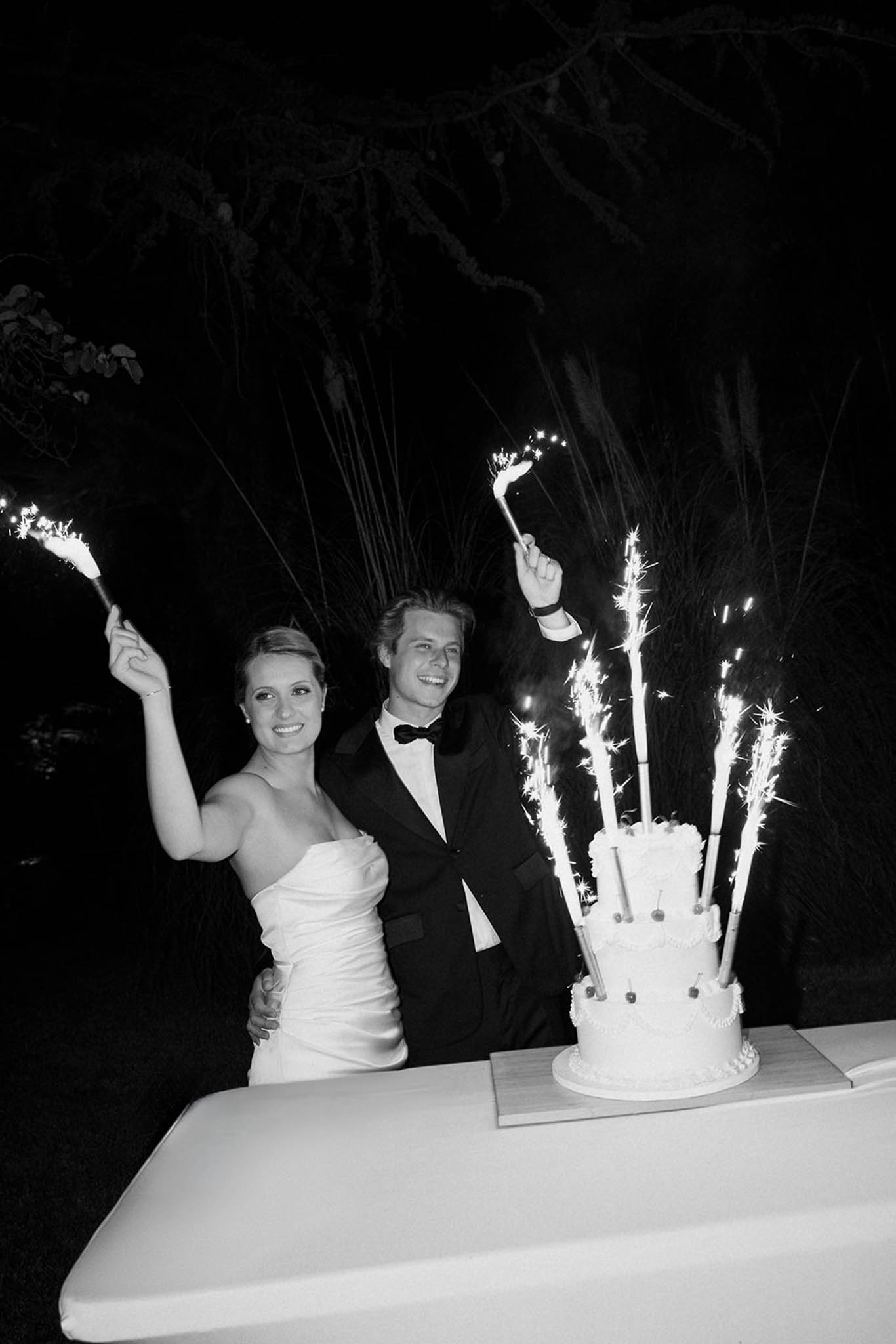 Bride and groom cutting two-tier white wedding cake at night outdoors, both holding lit sparklers, black and white photo