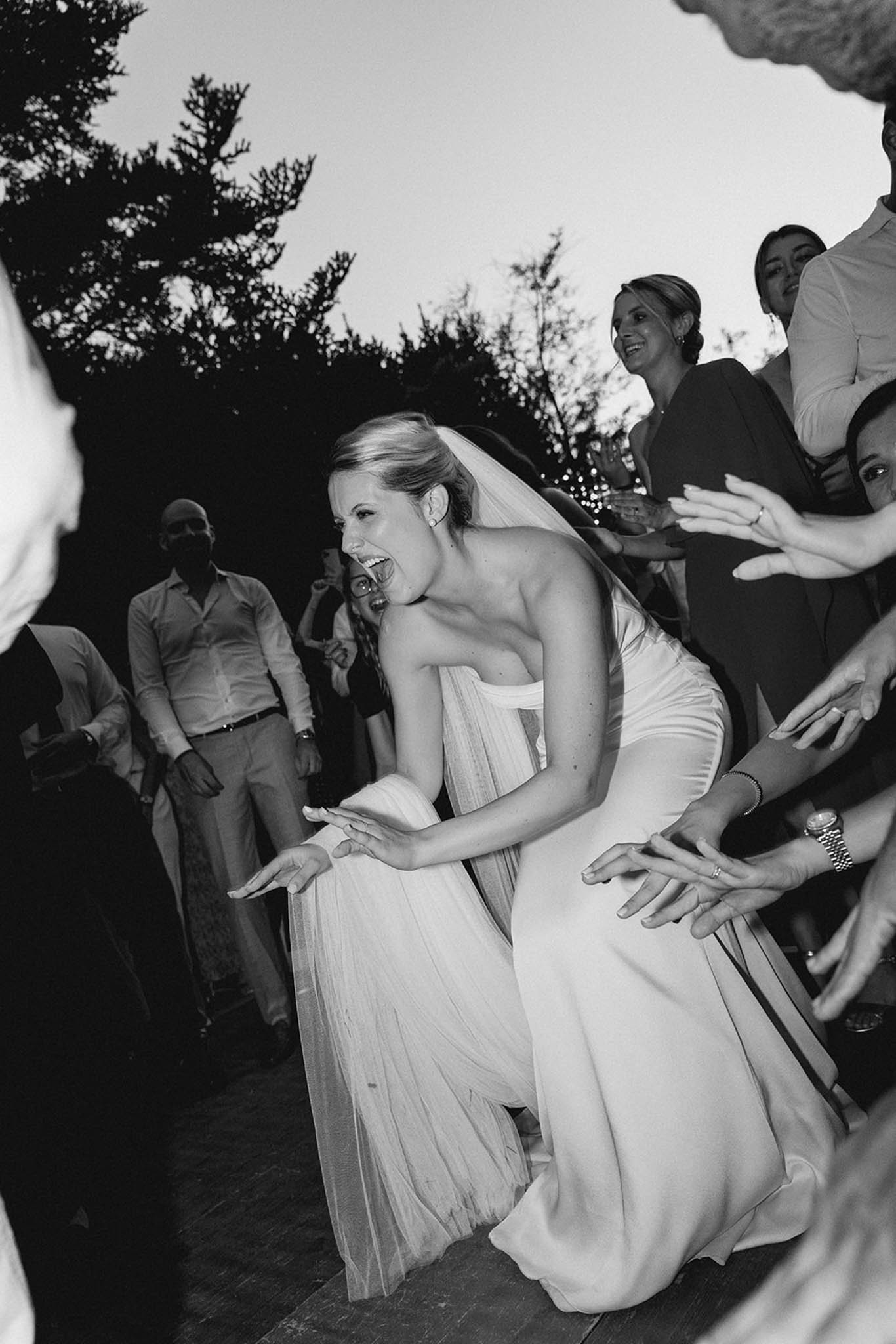 Bride in strapless pleated gown dancing joyfully with guests during outdoor reception in black and white
