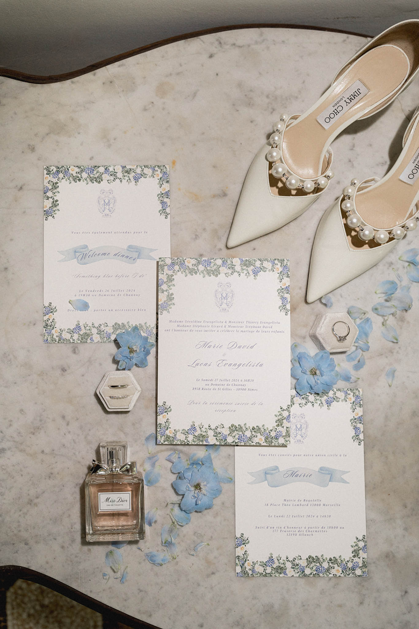 Flat lay on marble: cream invitations with periwinkle floral borders, ivory pearl-embellished heels, ring dish and Miss Dior perfume.