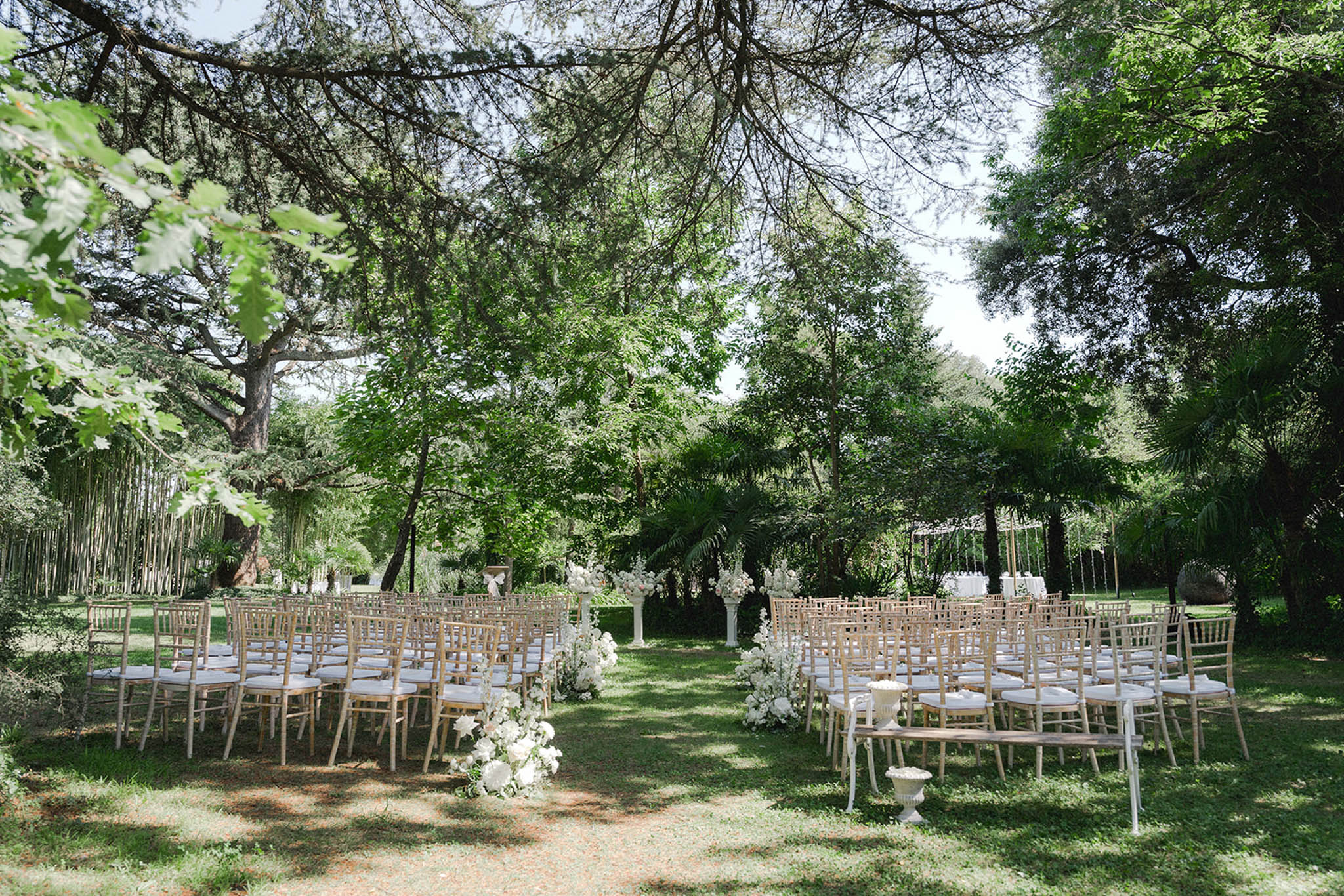 Outdoor ceremony seating under mature tree canopy with wood chairs and white floral aisle markers