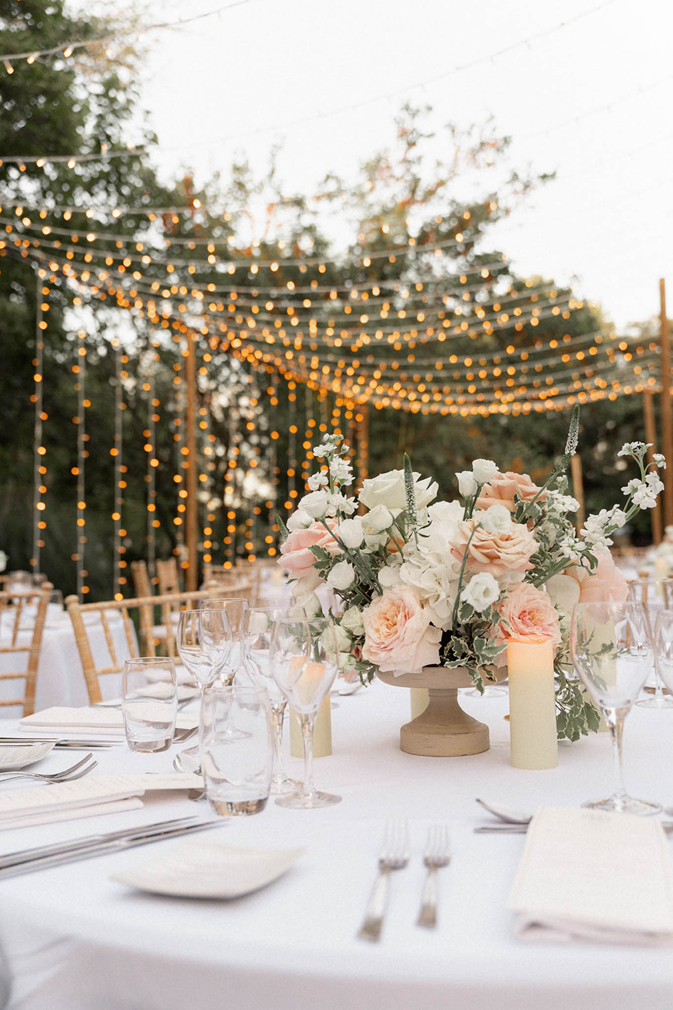 Outdoor reception table with peach and white rose centerpiece in a taupe pedestal vase, string lights in wooden pergola behind.