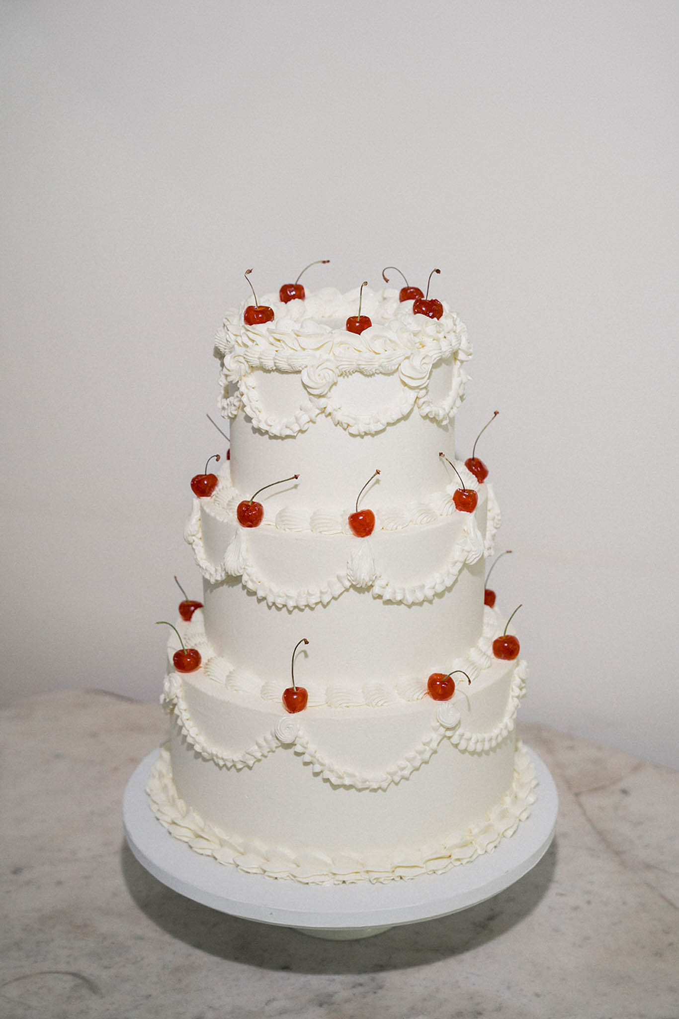 Four-tiered ivory buttercream wedding cake with scalloped piping and red maraschino cherry decorations on marble surface
