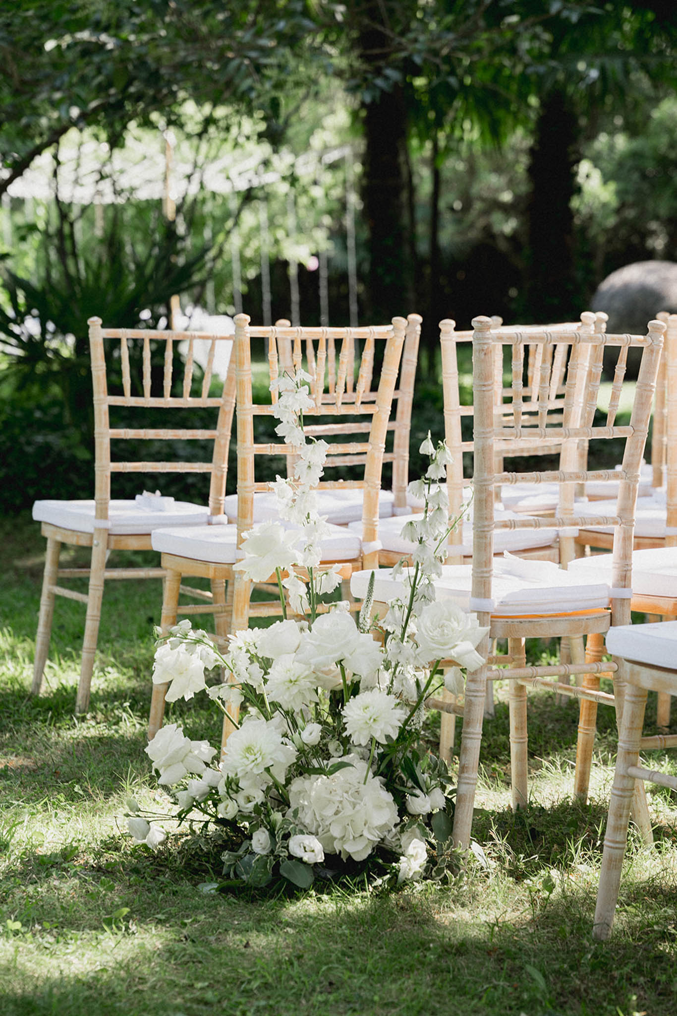 Ceremony aisle lined with Chiavari chairs and white floral arrangements of roses, hydrangeas and delphiniums on a garden lawn.