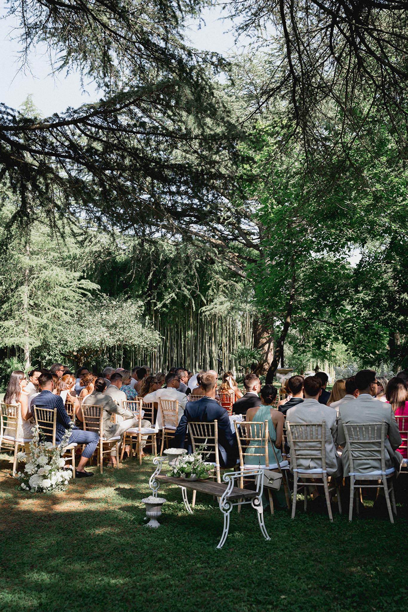 Outdoor ceremony under tree canopy with macramÃ© backdrop, 60-80 guests on Chiavari chairs and white florals