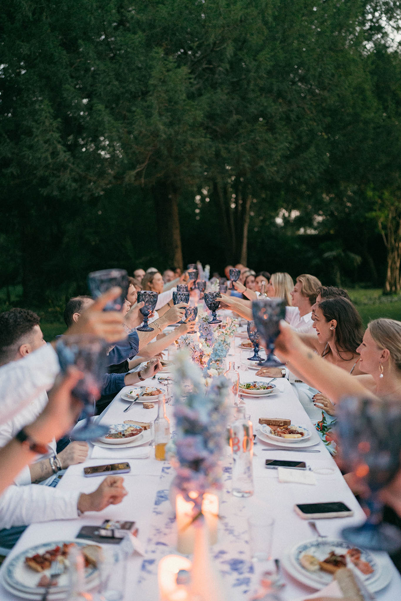 Guests seated at long outdoor dinner table beneath tall trees with purple floral centerpieces and candles at dusk