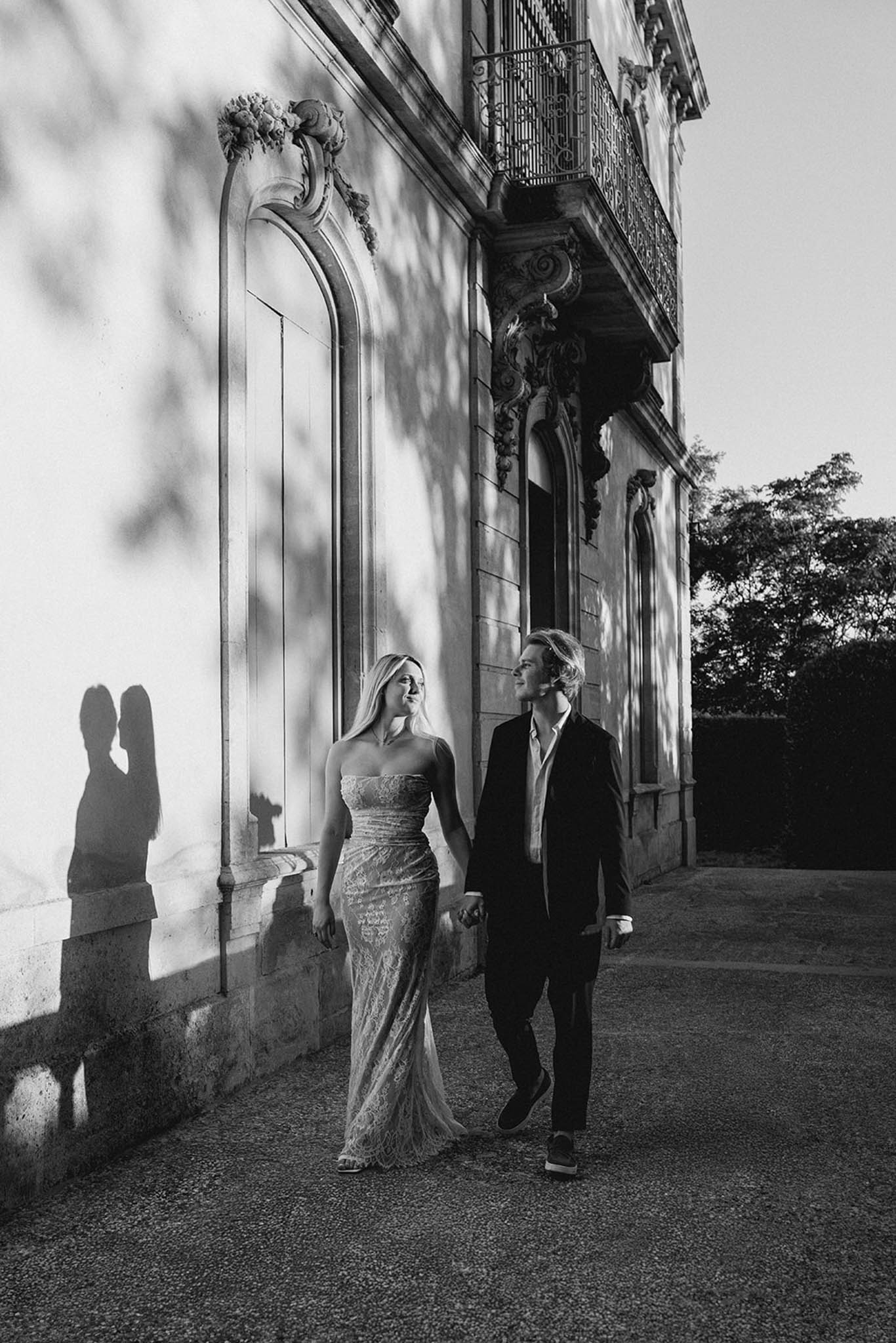 Black and white portrait of bride and groom walking along neoclassical stone building, dramatic shadows cast on facade