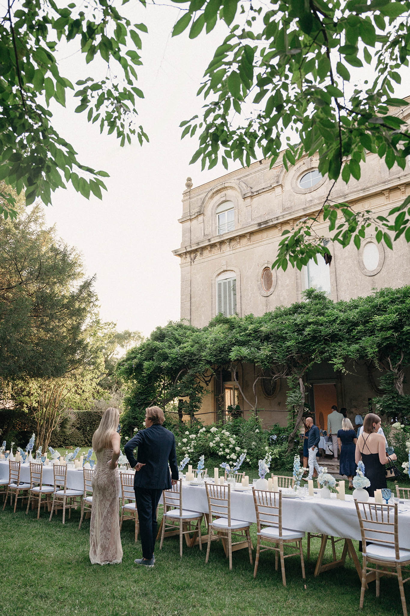 Outdoor reception tables with ivory linens and gold chairs beneath foliage in front of a historic stone palazzo.