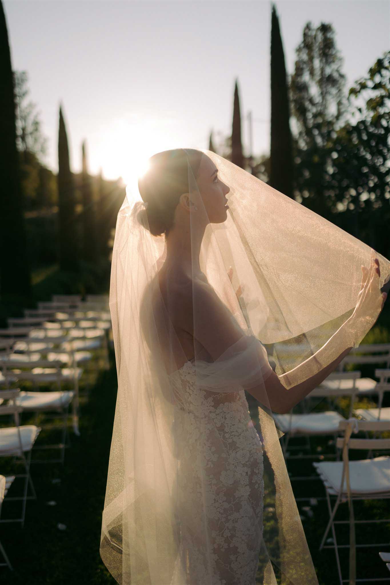 Bride in profile with long tulle veil billowing in the wind in a garden lined with cypress trees