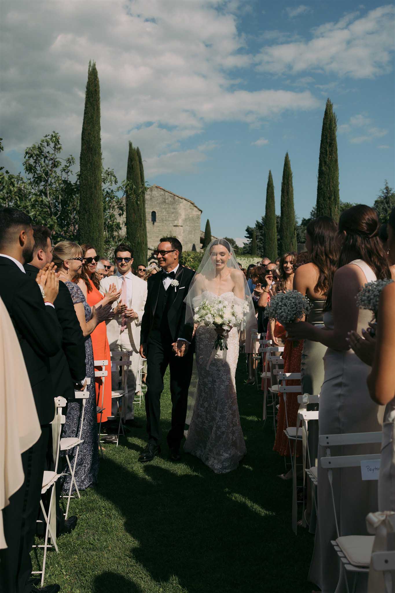 Bride and groom walk back down aisle past applauding guests after ceremony, framed by Italian cypress trees