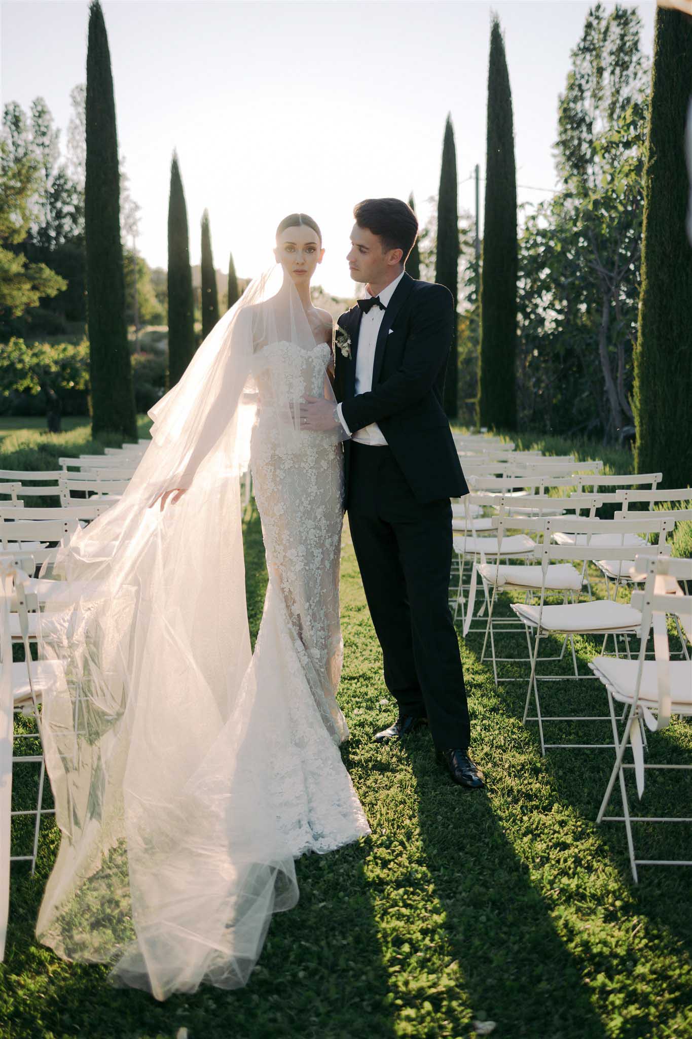 Bride in ivory beaded gown and groom in black tuxedo walk the aisle flanked by white folding chairs and tall cypress trees