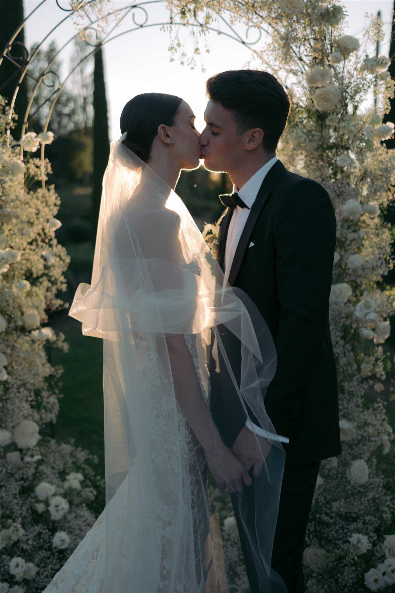 Bride and groom share first kiss under wrought-iron arch decorated with ivory roses and baby's breath