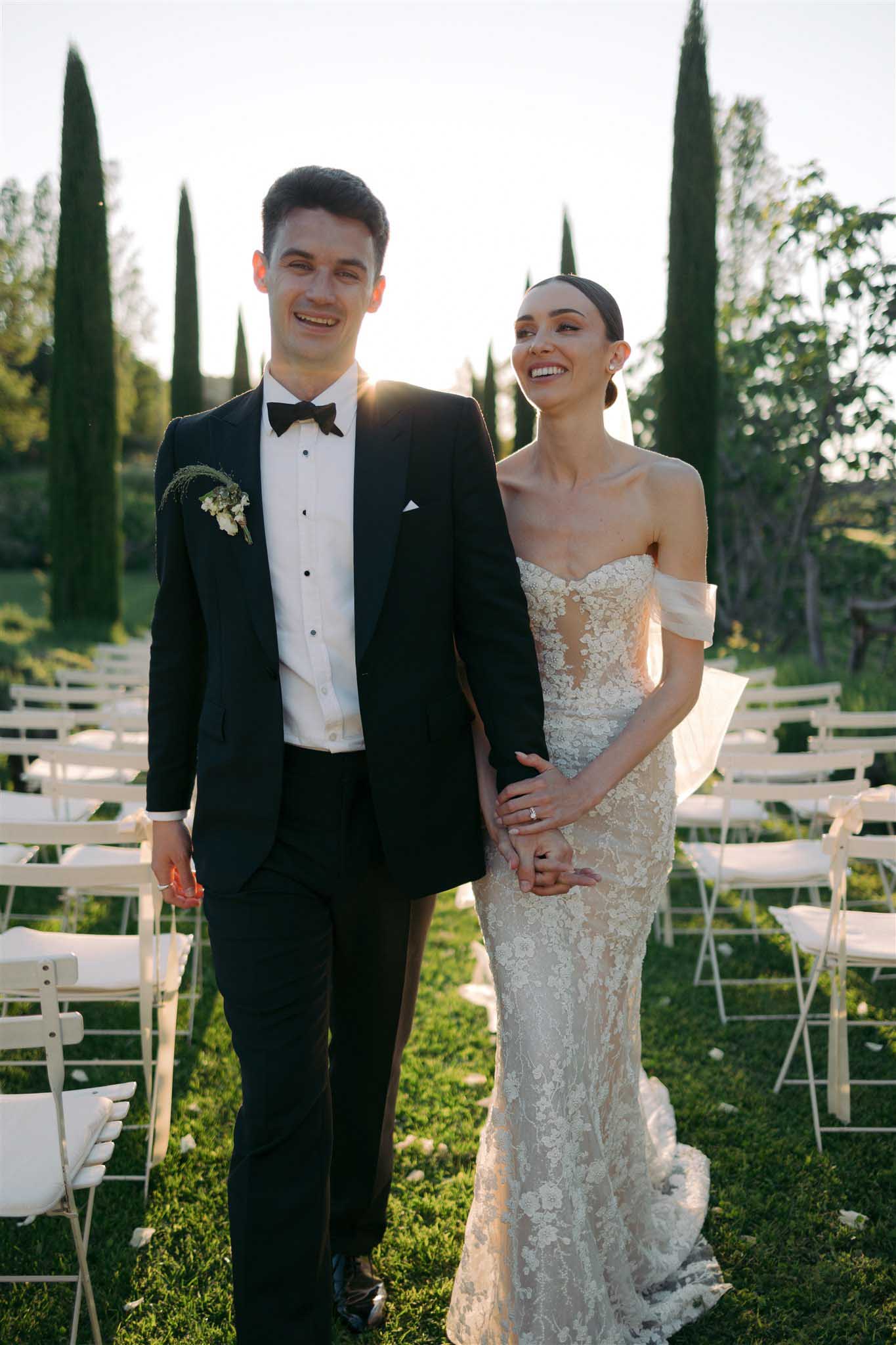 Bride and groom walking hand-in-hand down the aisle after the ceremony, guests in white chairs, cypress trees in background.