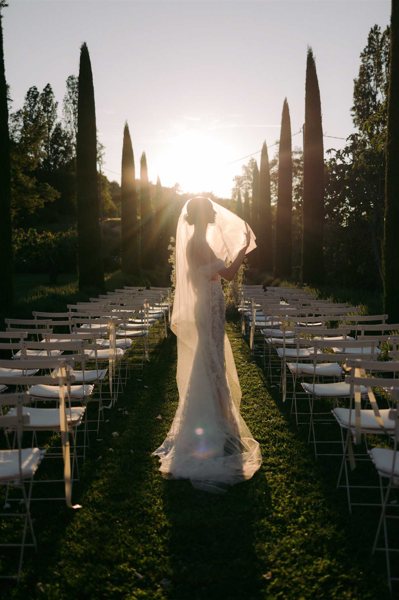 Bride in ivory lace gown with long veil stands alone down cypress-lined ceremony aisle backlit by golden sunset