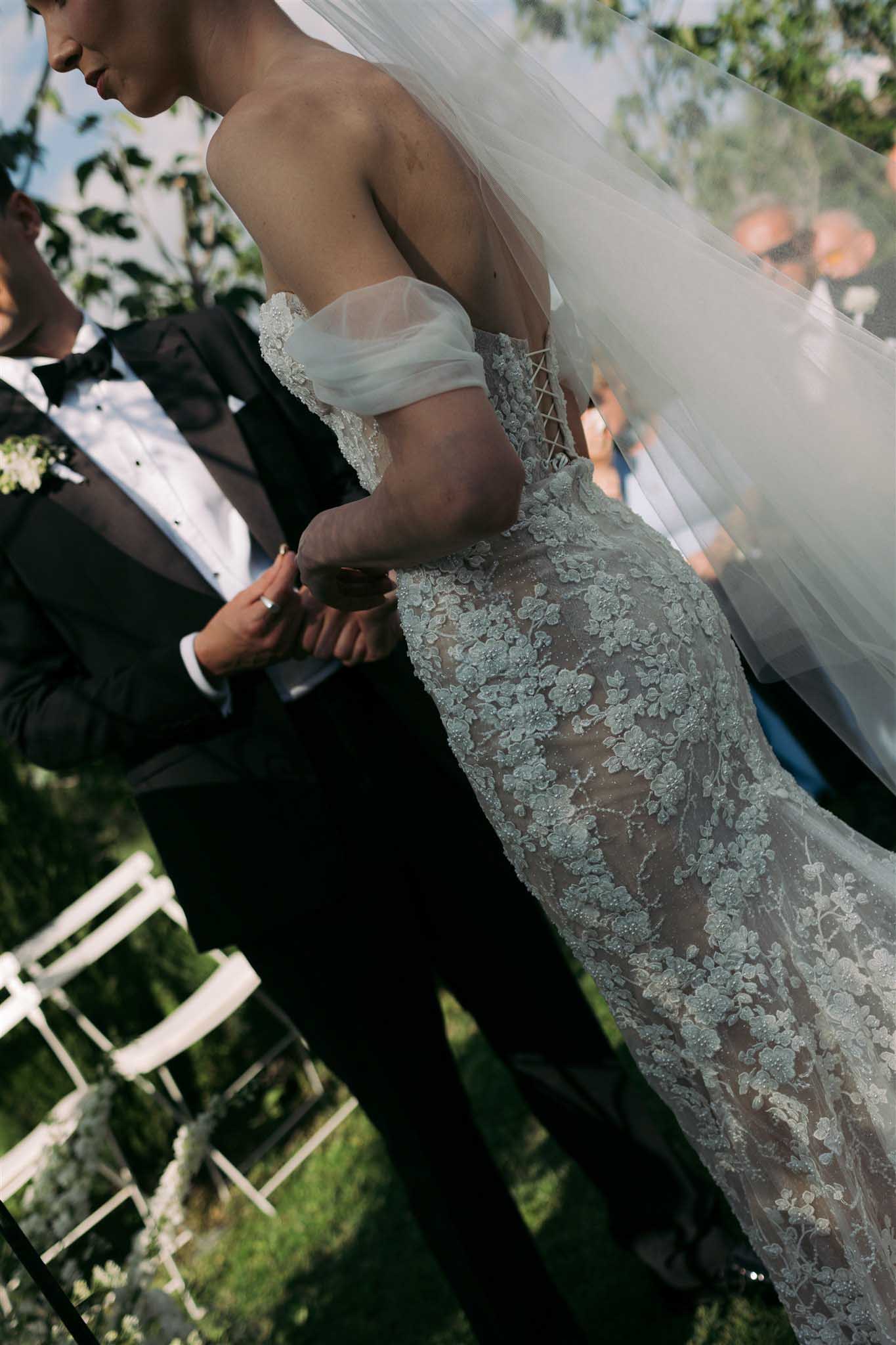 Bride in off-shoulder ivory lace gown with floral appliquÃ©s and voluminous veil during the outdoor ceremony, groom beside her.