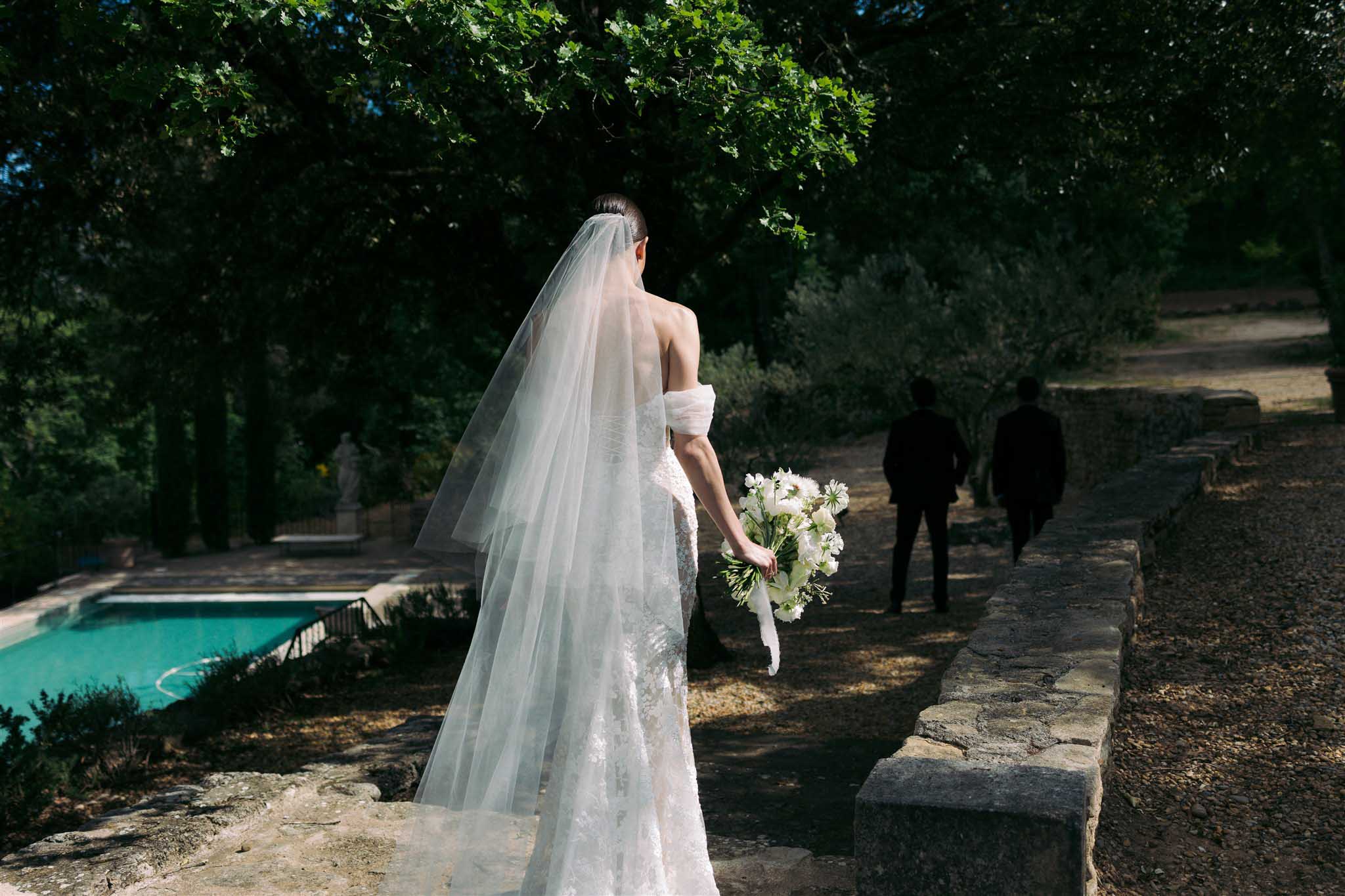 Bride in ivory lace gown from behind on garden path beside turquoise pool, long veil flowing with cypress trees