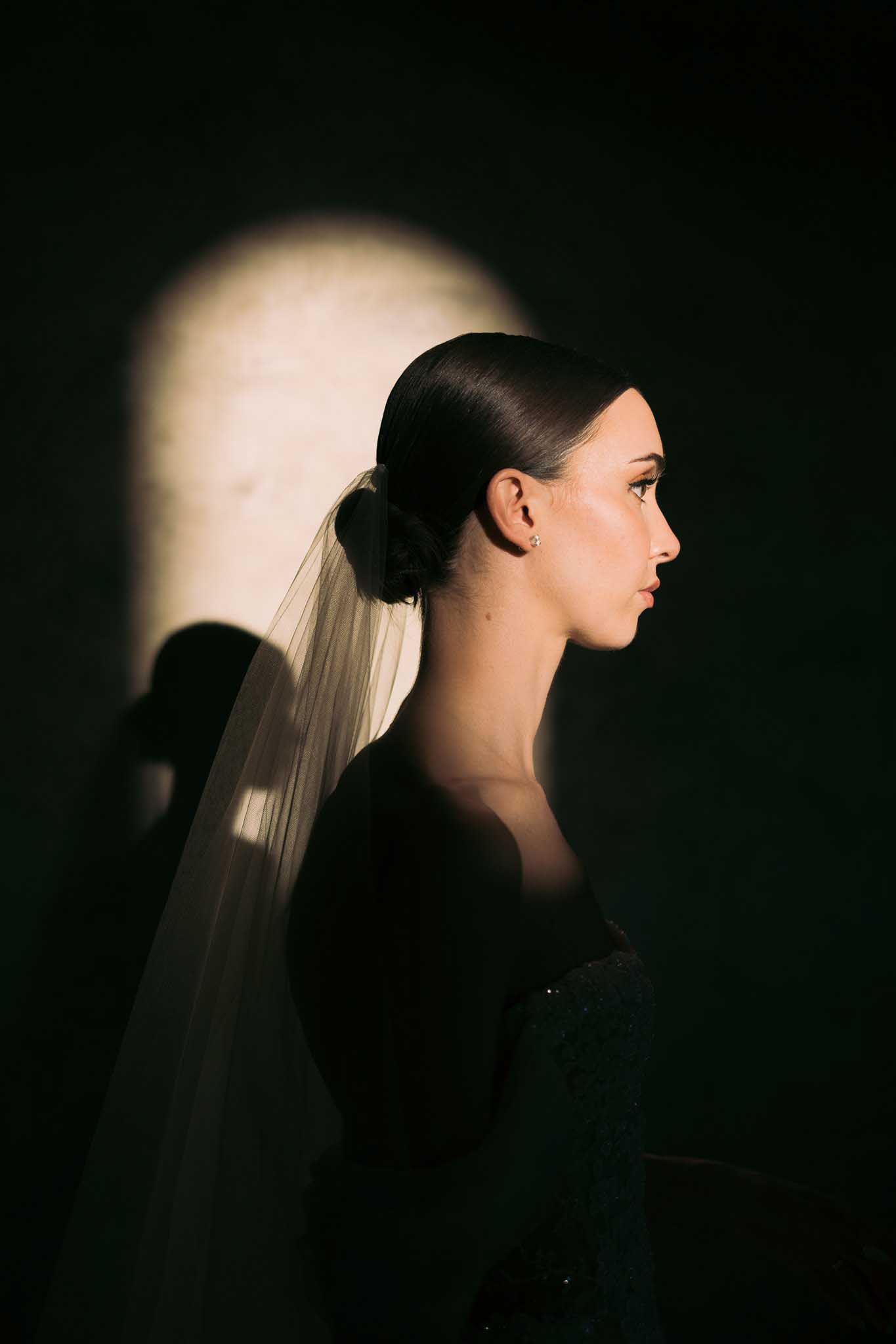 Bride in profile with sleek low bun and flowing ivory veil, dramatic directional lighting against dark background