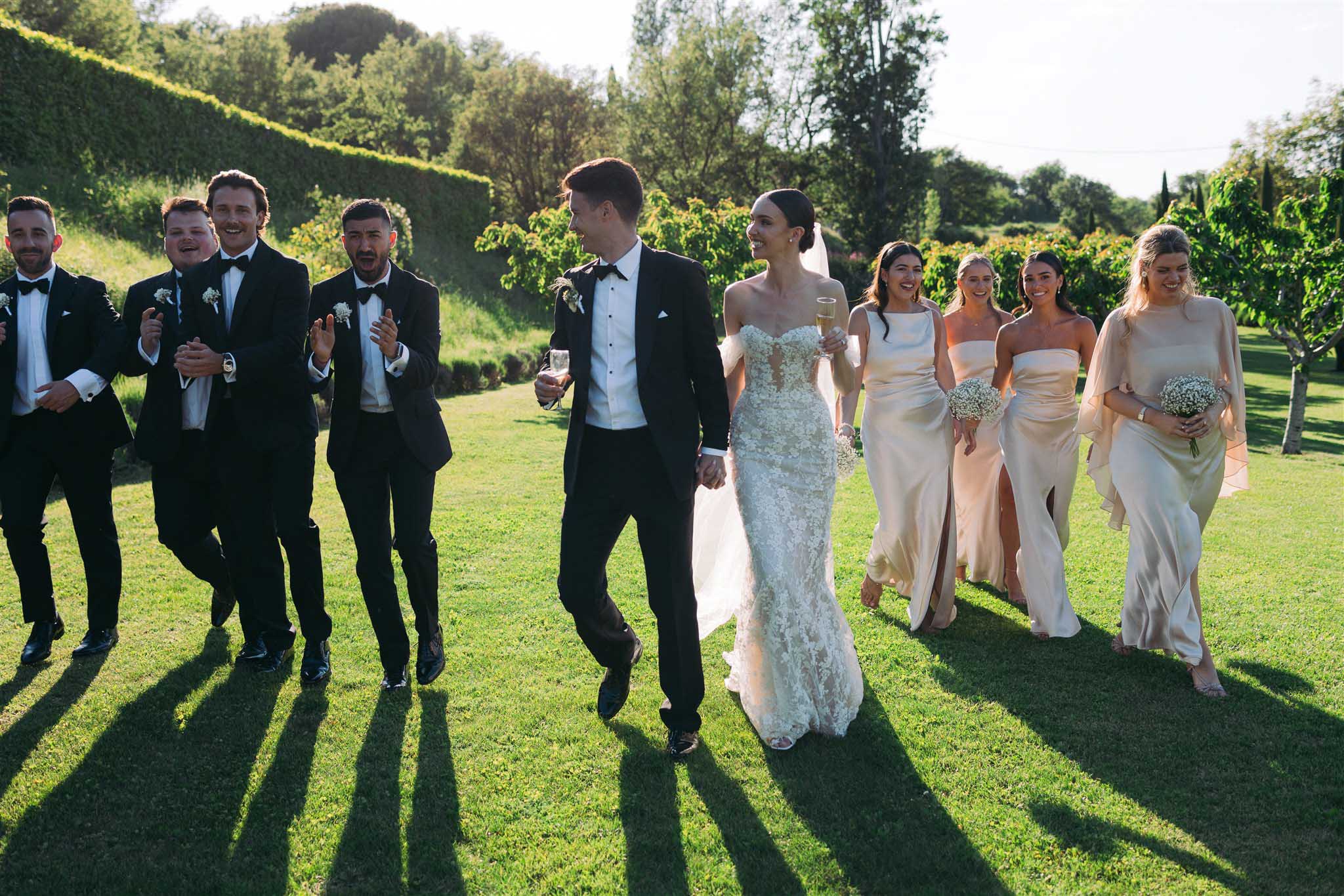 Full bridal party walking across a manicured garden; bride in lace gown flanked by groomsmen in tuxedos and bridesmaids in champagne gowns.