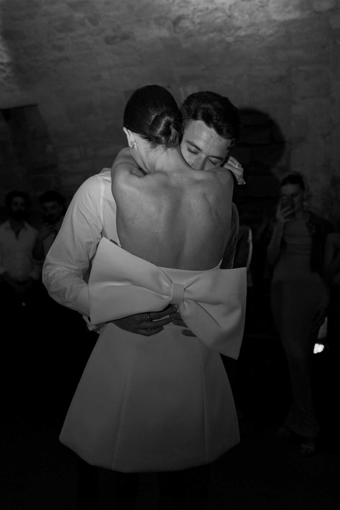 Groom embracing bride from behind during first dance, black and white, minimalist dress with bow back, guests behind