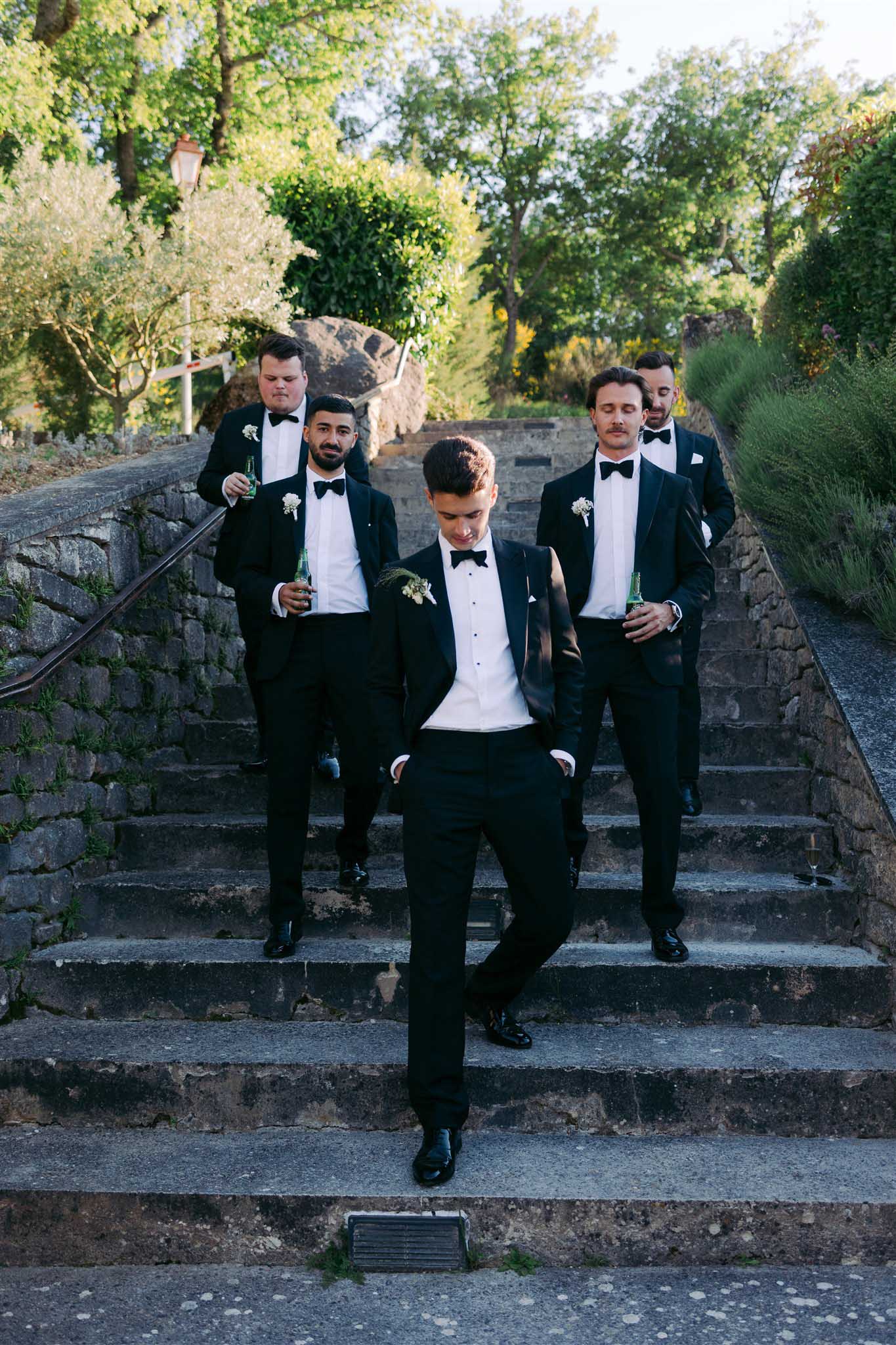 Groom and four groomsmen in black tuxedos posing on moss-covered stone staircase with champagne flutes