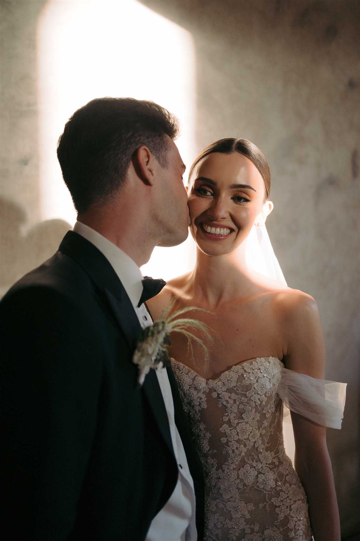 Groom kissing bride on the cheek in sunlight at Abbaye Saint-Eusebe photographed by Ayata