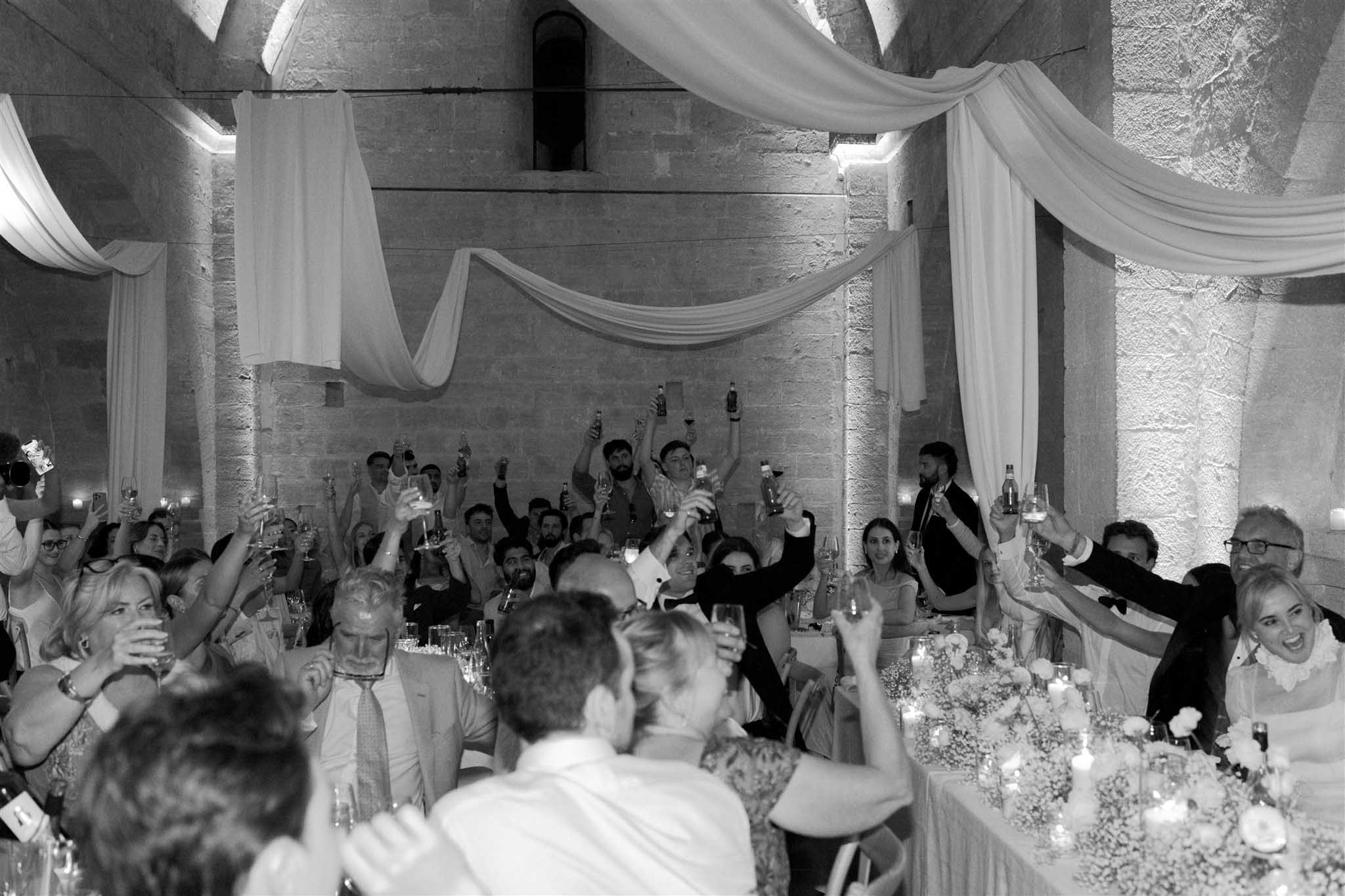 Black and white wide shot of guests raising glasses in toast at long tables inside stone-walled reception venue