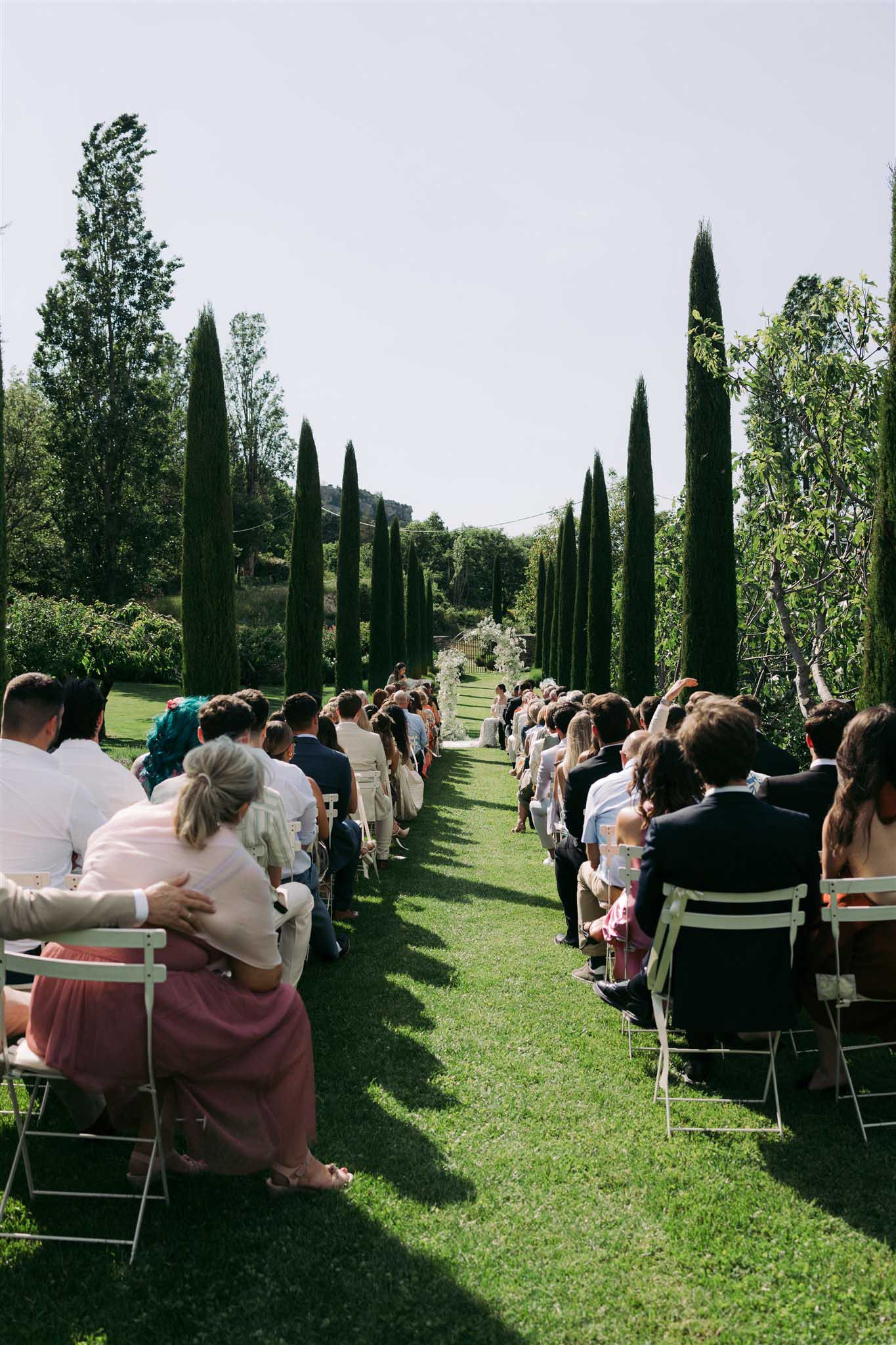 Wide shot of 80-100 guests in white chairs down long cypress-lined aisle at Mediterranean garden ceremony