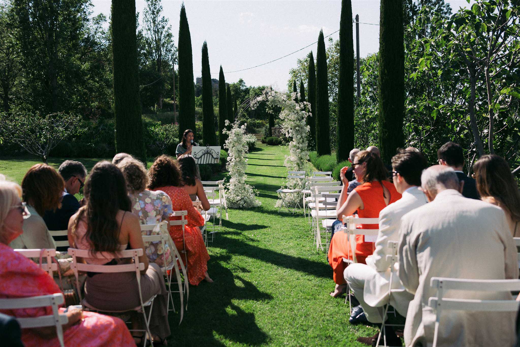 Outdoor garden ceremony with white floral arch, rows of guests on white chairs and tall cypress trees