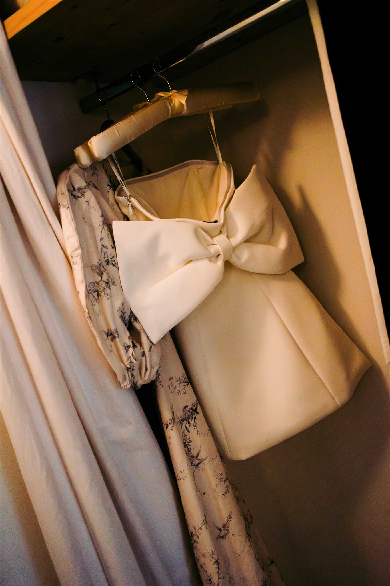 Ivory wedding dress with silk bow and pale yellow bridesmaid dress hanging beside cream floral-print guest dress