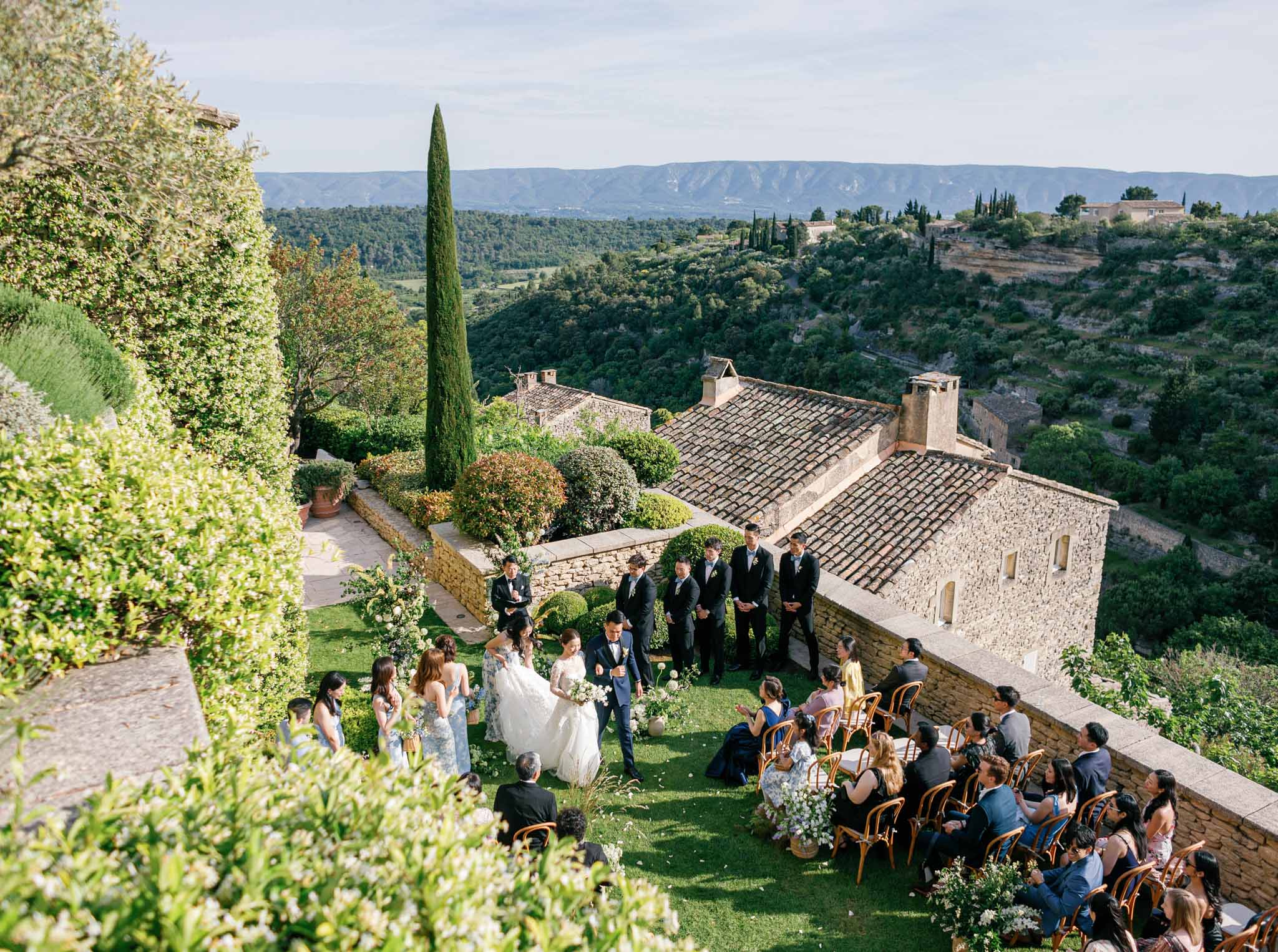 Aerial view of outdoor ceremony on hillside with guests in wooden chairs overlooking a Mediterranean valley