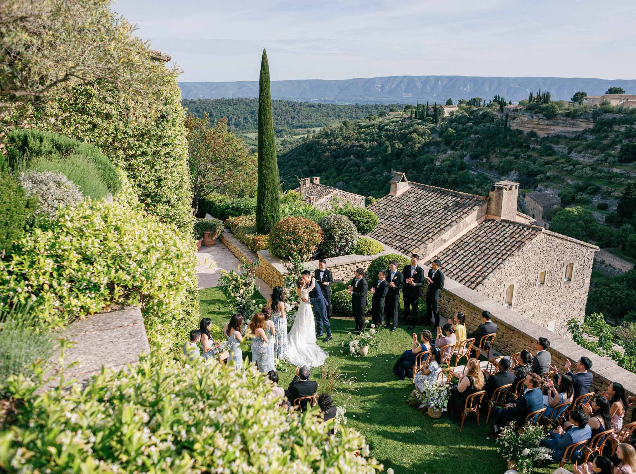 Aerial view of outdoor ceremony on lawn of Provence stone villa with guests on Chiavari chairs and floral arch