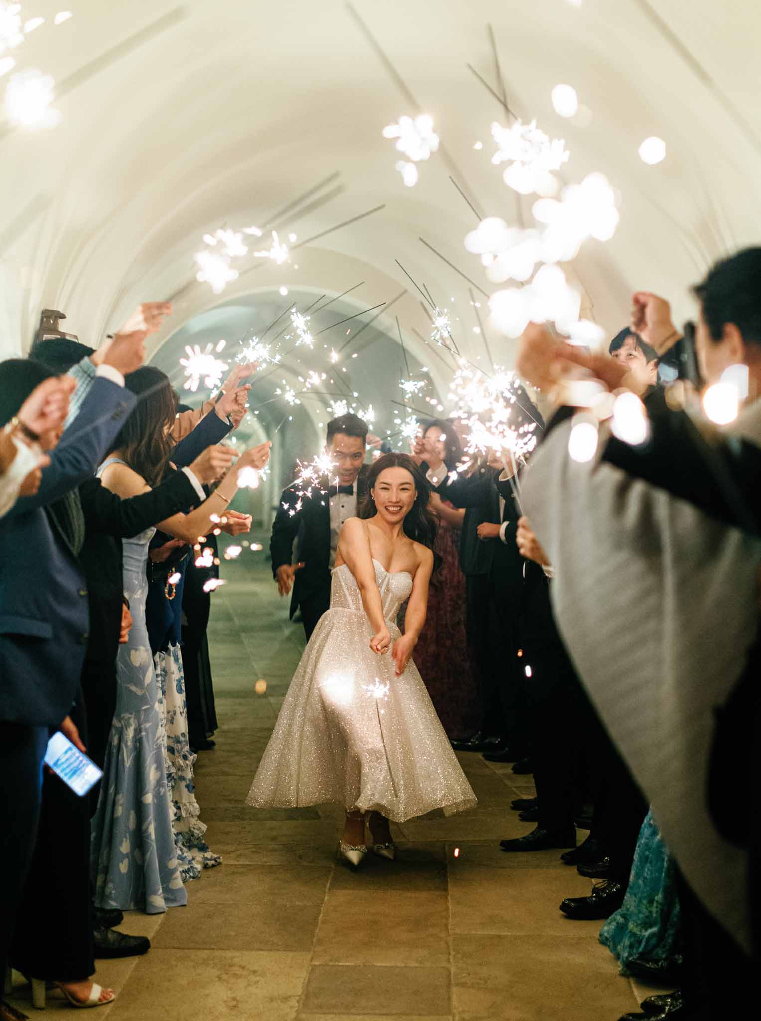 Bride in champagne-gold sequined gown and groom walking through a sparkler tunnel inside an arched stone corridor.