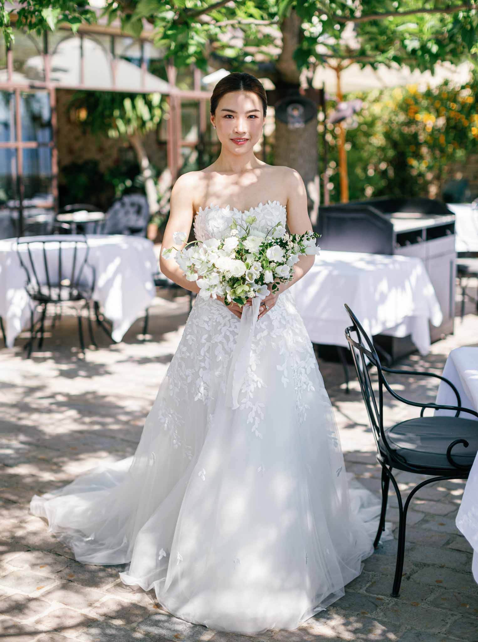 Full-length bridal portrait with white rose bouquet in outdoor courtyard with round reception tables behind