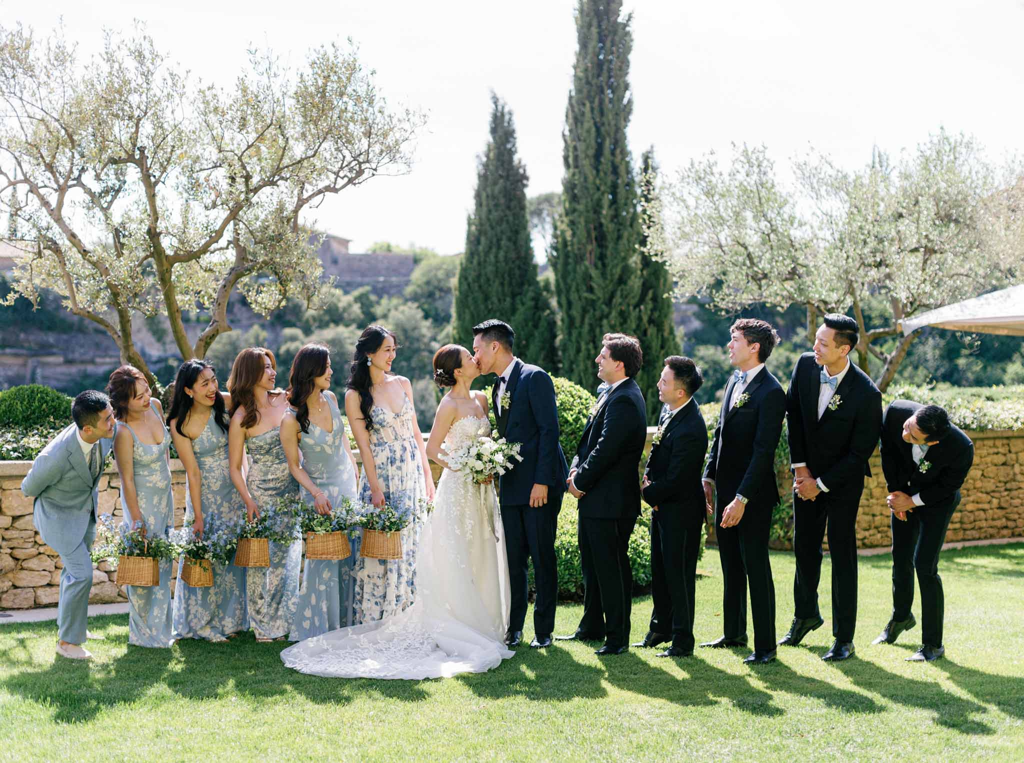 Bridal party lined up in garden with olive trees, bridesmaids in blue floral dresses and groomsmen in navy suits flanking kissing couple