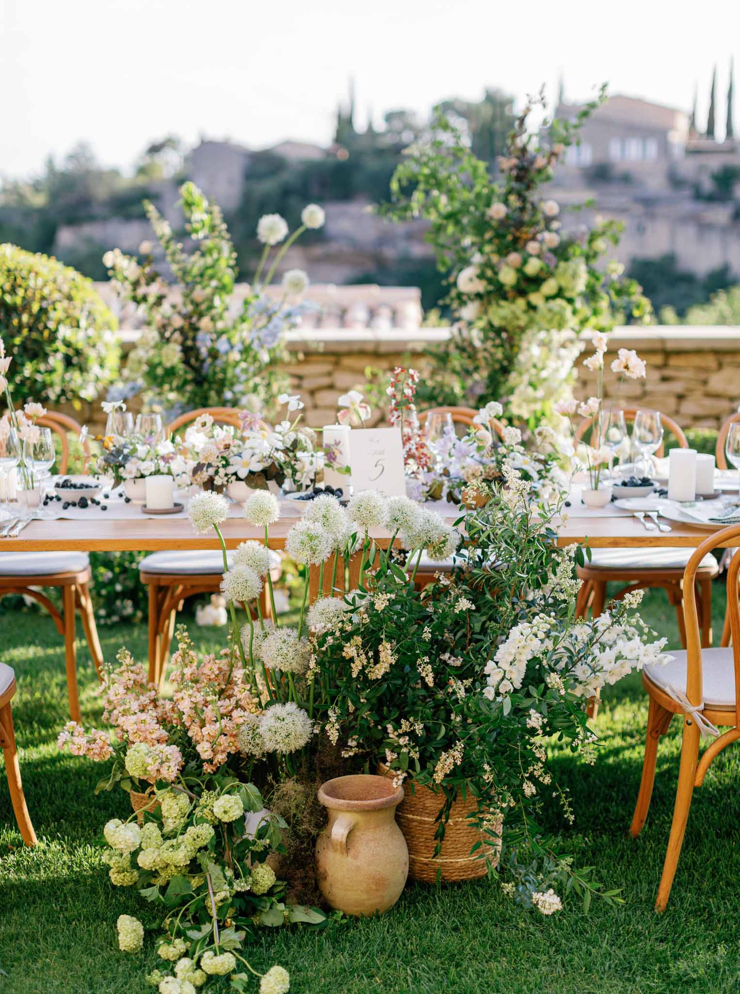 Outdoor reception table on lawn with lush white and blush floral arrangements in terracotta vessels overlooking hillside