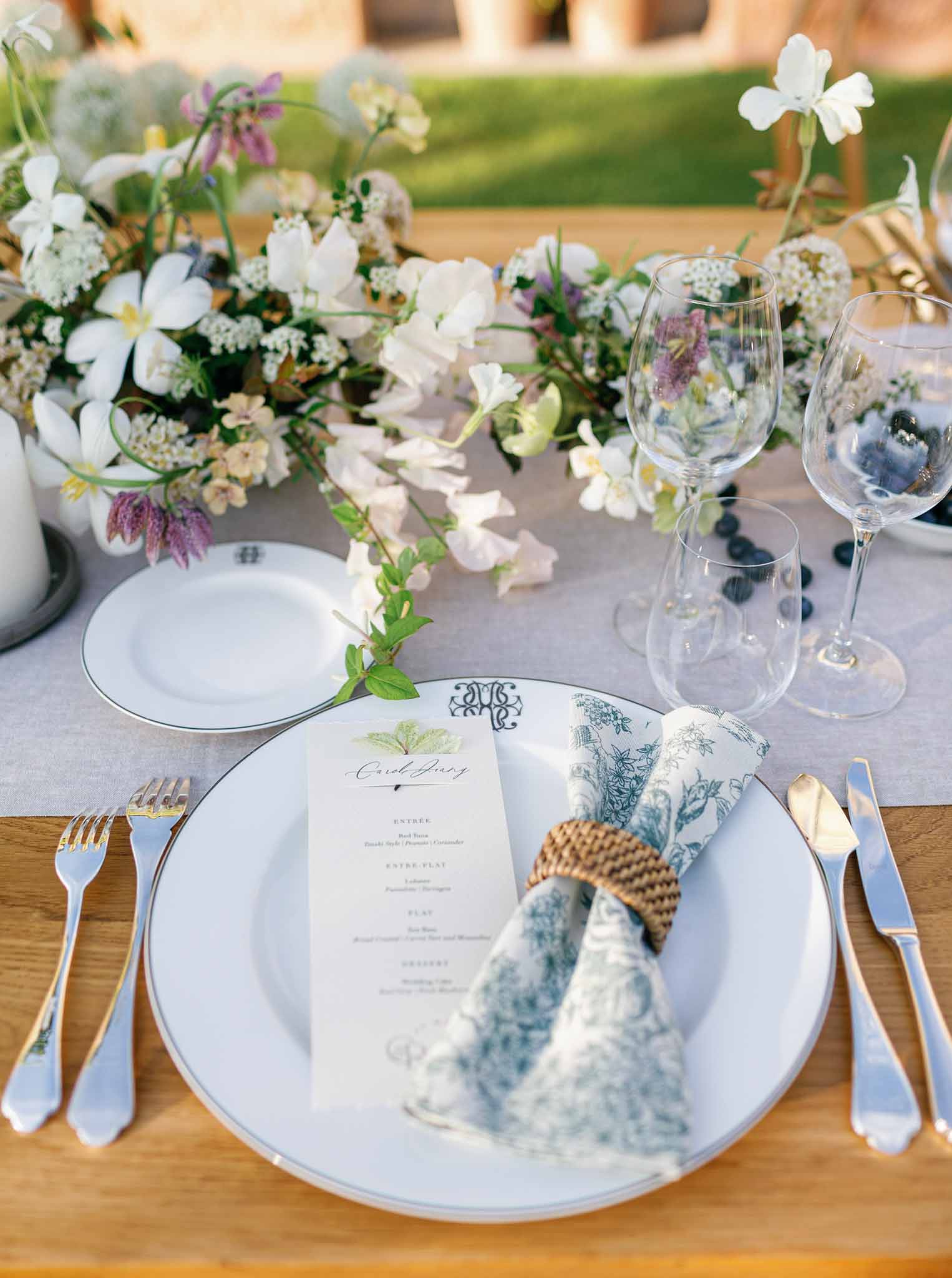 Place setting with cream menu card, blue botanical napkin, gold flatware, and white florals