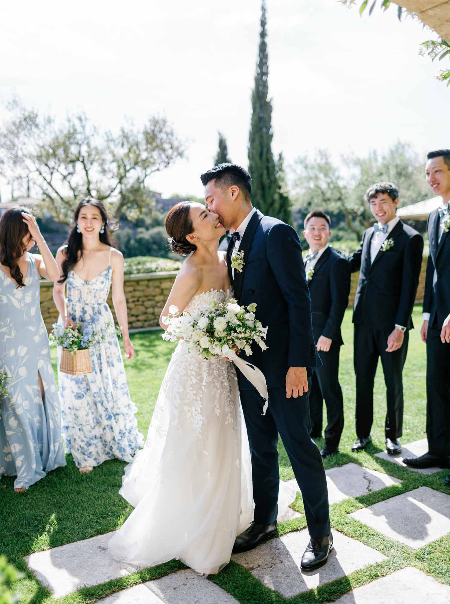 Bride and groom kiss during outdoor garden ceremony surrounded by bridal party with cypress trees and olive groves beyond
