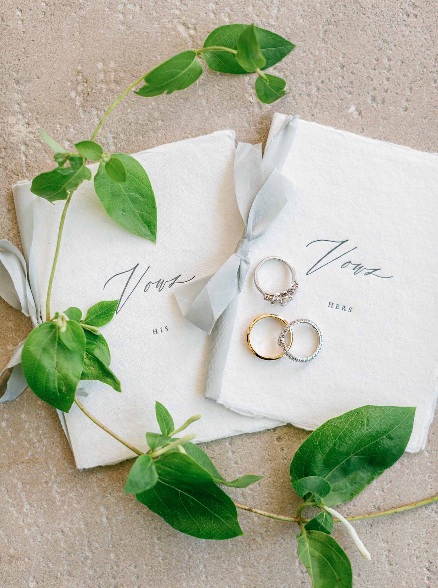 Flat lay of wedding rings, his and hers vow books with gray ribbon, and eucalyptus sprigs on stone surface