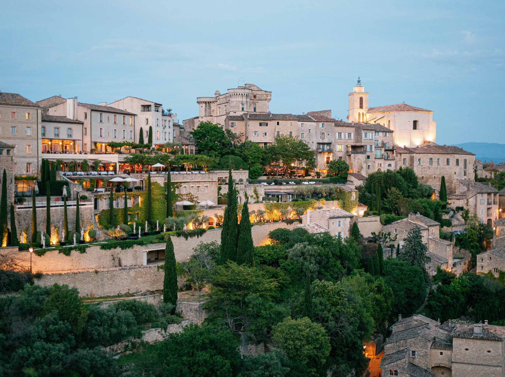ProvenÃ§al hilltop stone village at twilight with warm golden lighting, church bell tower, and cypress-lined terraces
