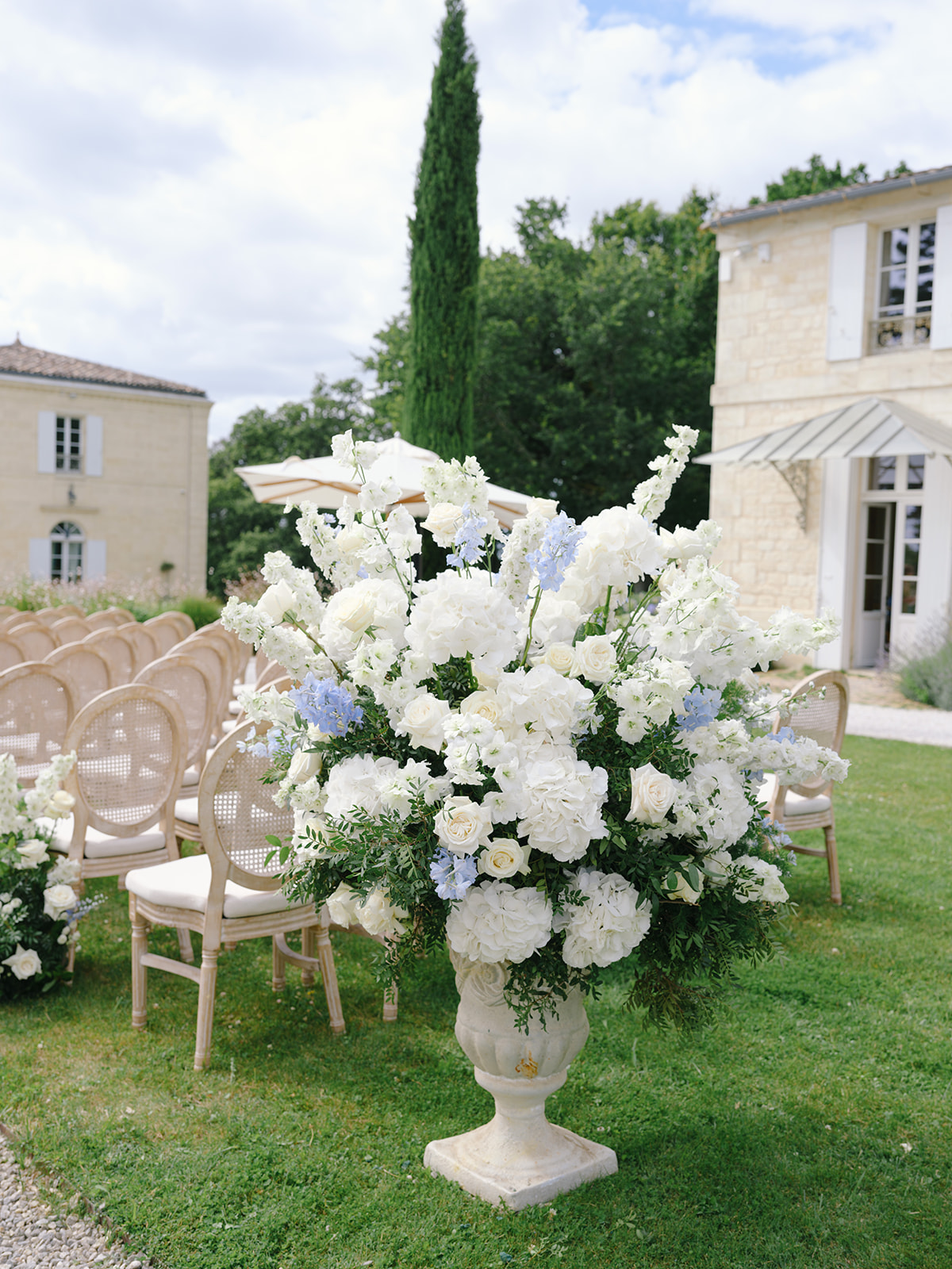 Large floral arrangement of white peonies, roses, blush roses and delphiniums in a stone urn beside ceremony chairs on a lawn.
